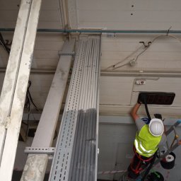 Lavador Łukasz Biazik - Pracownik w kasku i kamizelce odblaskowej na drabinie w Łodzi, czyści przemysłową instalację pod sufitem hali, używając odkurzacza przemysłowego.