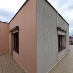 Lavador Łukasz Biazik - Dwukolorowa elewacja domu z oknami zabezpieczonymi roletami zewnętrznymi, widoczna z perspektywy tarasu wyłożonego płytkami.