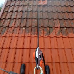 Lavador Łukasz Biazik - Częściowo wyczyszczony dach z czerwonej dachówki, widoczne buty osoby czyszczącej i lina zabezpieczająca z osprzętem.