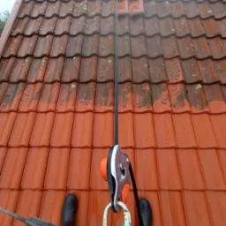 Częściowo wyczyszczony dach z czerwonej dachówki, widoczne buty osoby czyszczącej i lina zabezpieczająca z osprzętem.