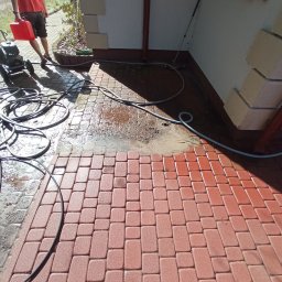 Lavador Łukasz Biazik - Czerwona kostka brukowa częściowo umyta, widoczna różnica w kolorze, wąż ciśnieniowy i myjka ciśnieniowa z czerwonym kanistrem w tle.