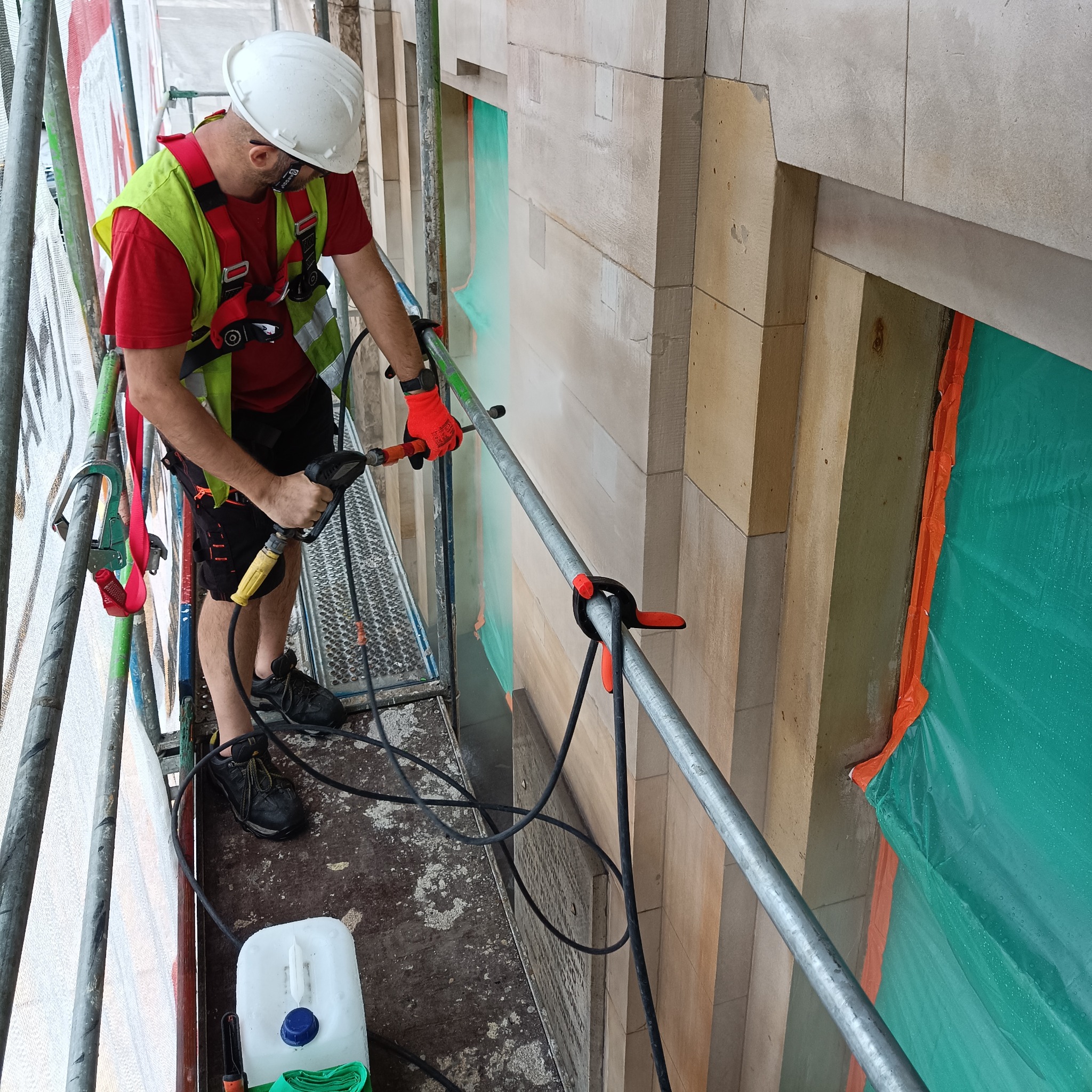 Pracownik w kasku i uprzęży na rusztowaniu czyści elewację kamienicy za pomocą myjki ciśnieniowej, zabezpieczając okna folią ochronną.