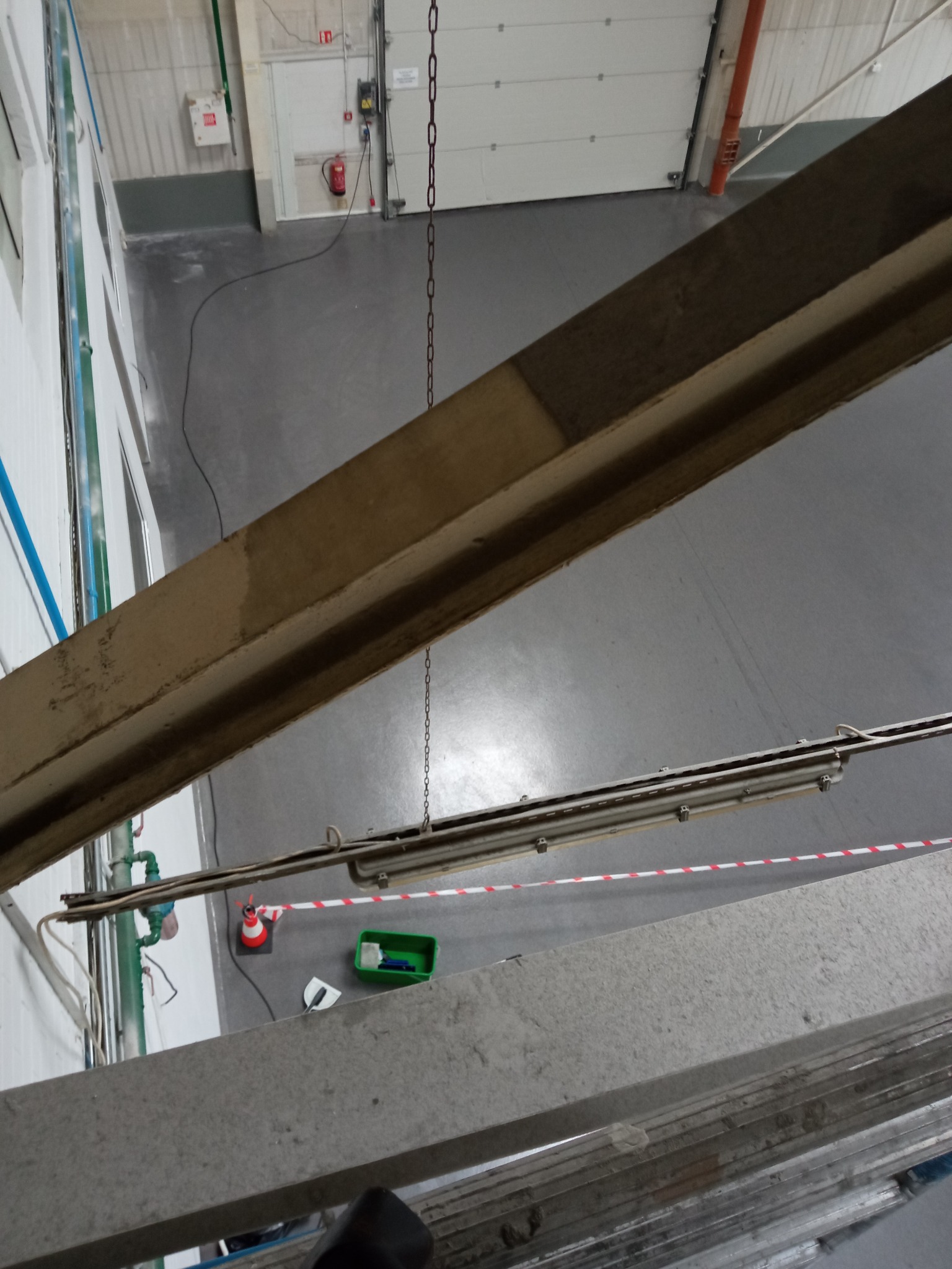 Widok z góry na przemysłową halę z betonową konstrukcją, lampą, taśmą ostrzegawczą, stożkiem i narzędziami sprzątającymi, sugerujący prace porządkowe.