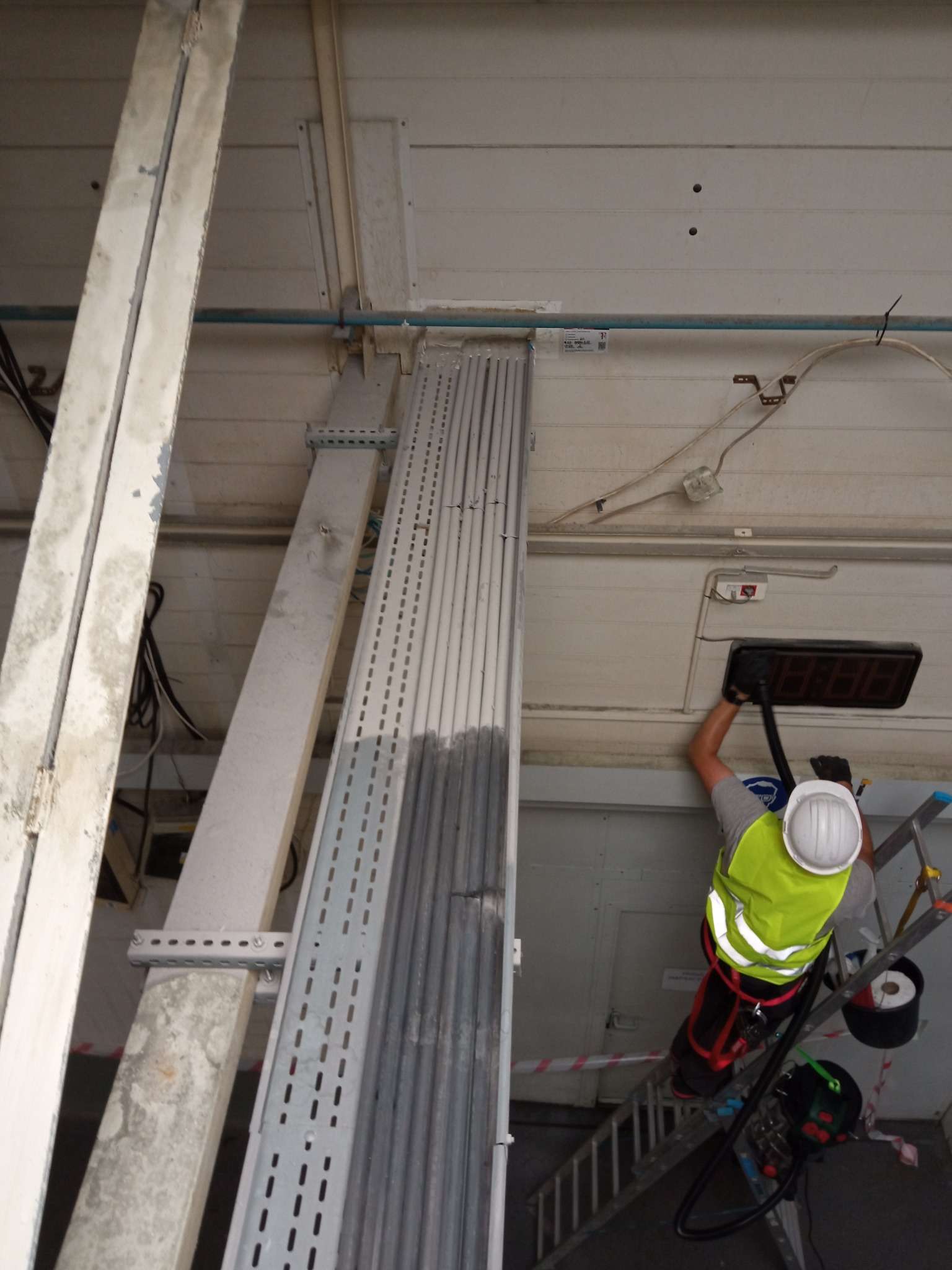 Pracownik w kasku i kamizelce odblaskowej na drabinie w Łodzi, czyści przemysłową instalację pod sufitem hali, używając odkurzacza przemysłowego.