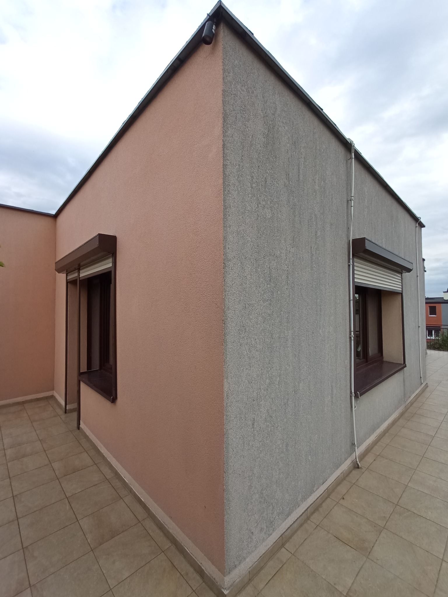 Dwukolorowa elewacja domu z oknami zabezpieczonymi roletami zewnętrznymi, widoczna z perspektywy tarasu wyłożonego płytkami.