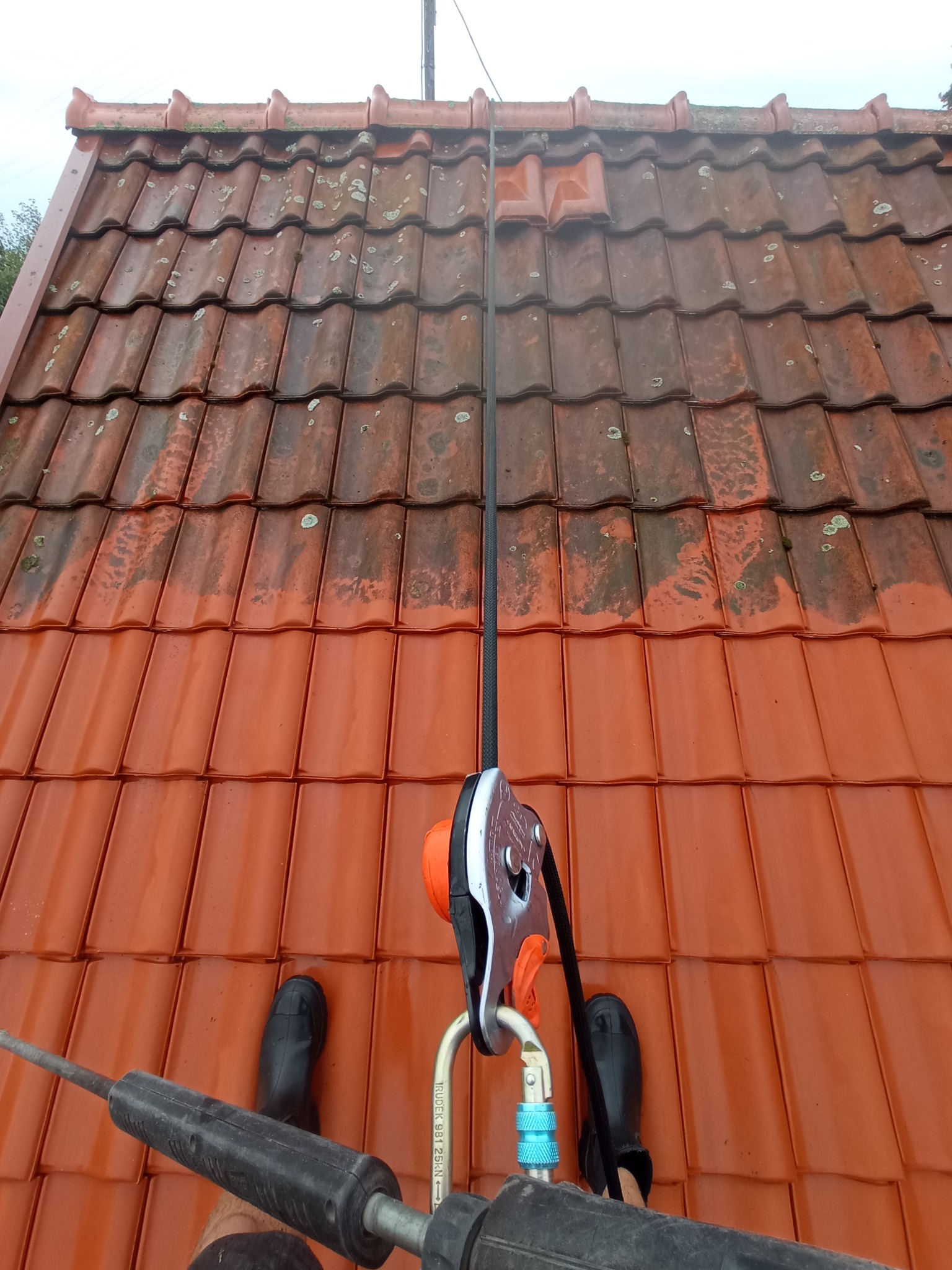 Częściowo wyczyszczony dach z czerwonej dachówki, widoczne buty osoby czyszczącej i lina zabezpieczająca z osprzętem.