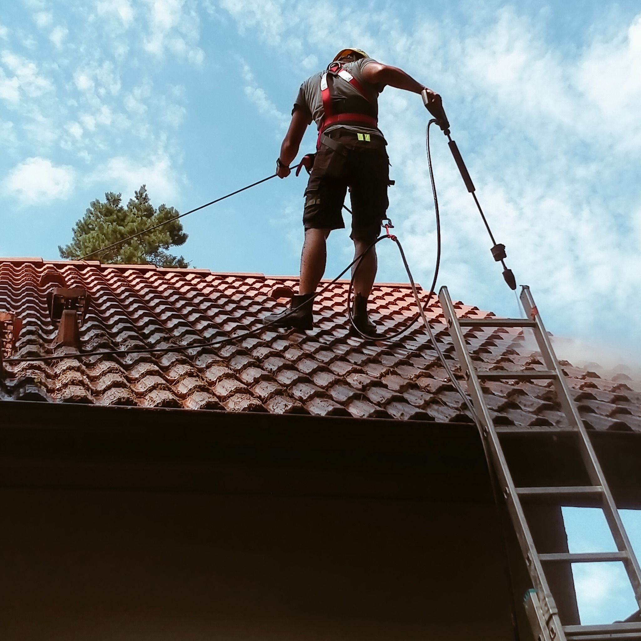 Pracownik w uprzęży bezpieczeństwa czyści dachówkę za pomocą myjki ciśnieniowej, stojąc na drabinie, na tle błękitnego nieba z chmurami.