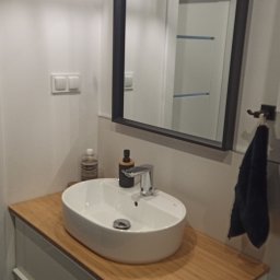 KRZYSZTOF KUŹNIK - Nowoczesna łazienka z owalną umywalką nablatową, drewnianym blatem, czarnym lustrem w ramie i designerską lampą sufitową, uzupełniona o dozownik mydła i ręcznik.