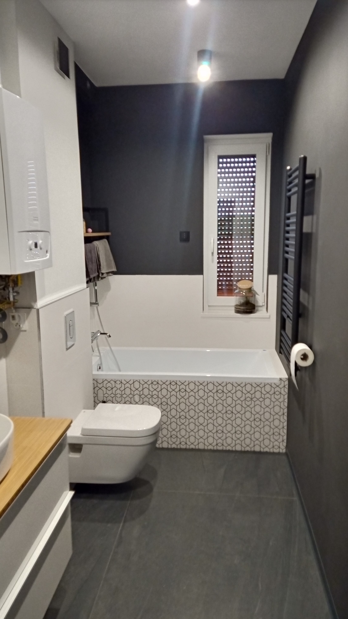 Wykończona łazienka w odcieniach szarości i bieli z wanną obudowaną płytkami w geometryczny wzór, wiszącą toaletą i grzejnikiem drabinkowym.