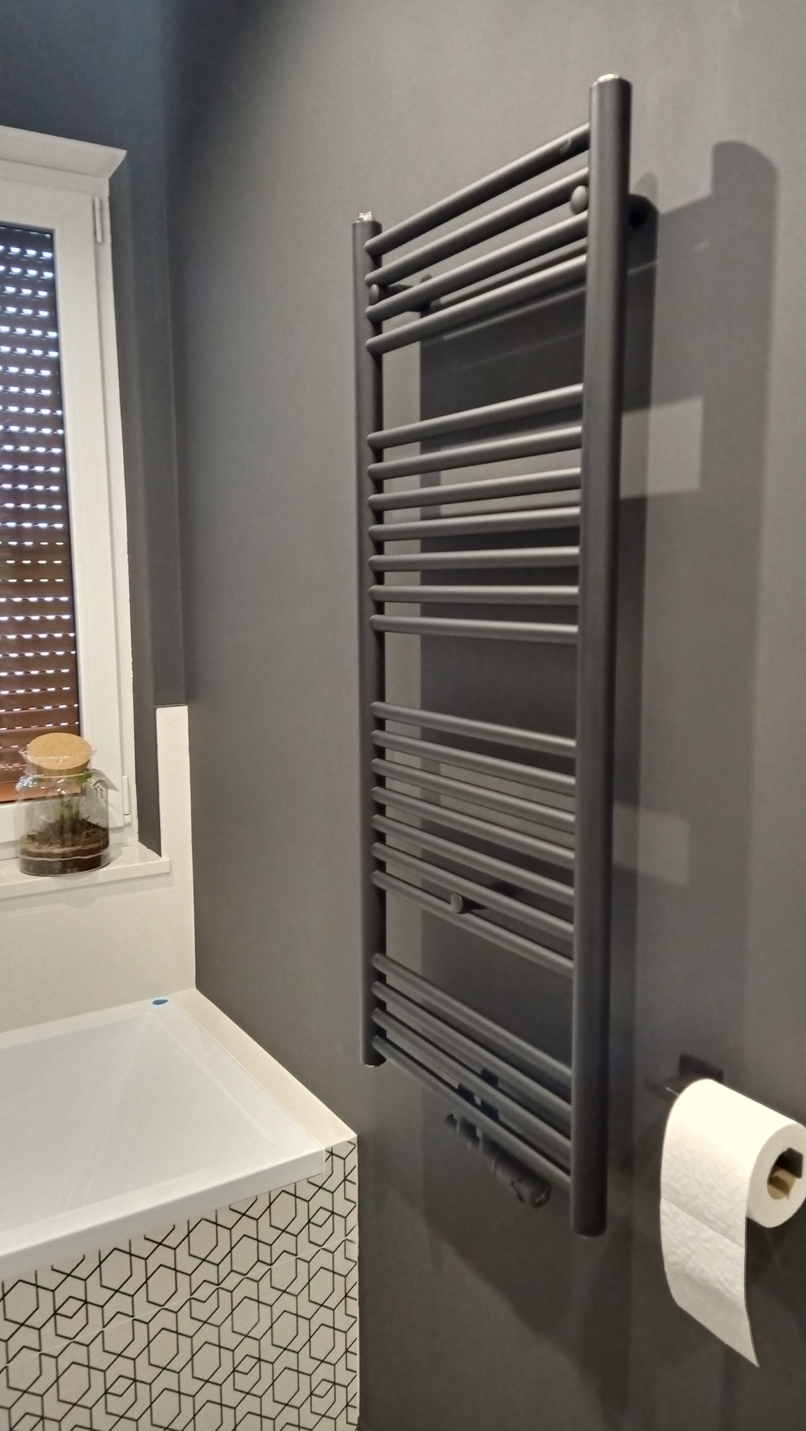 Szary grzejnik drabinkowy na ciemnej ścianie w łazience, obok toaleta i wanna z geometrycznym wzorem, na parapecie okna słoik z roślinami.