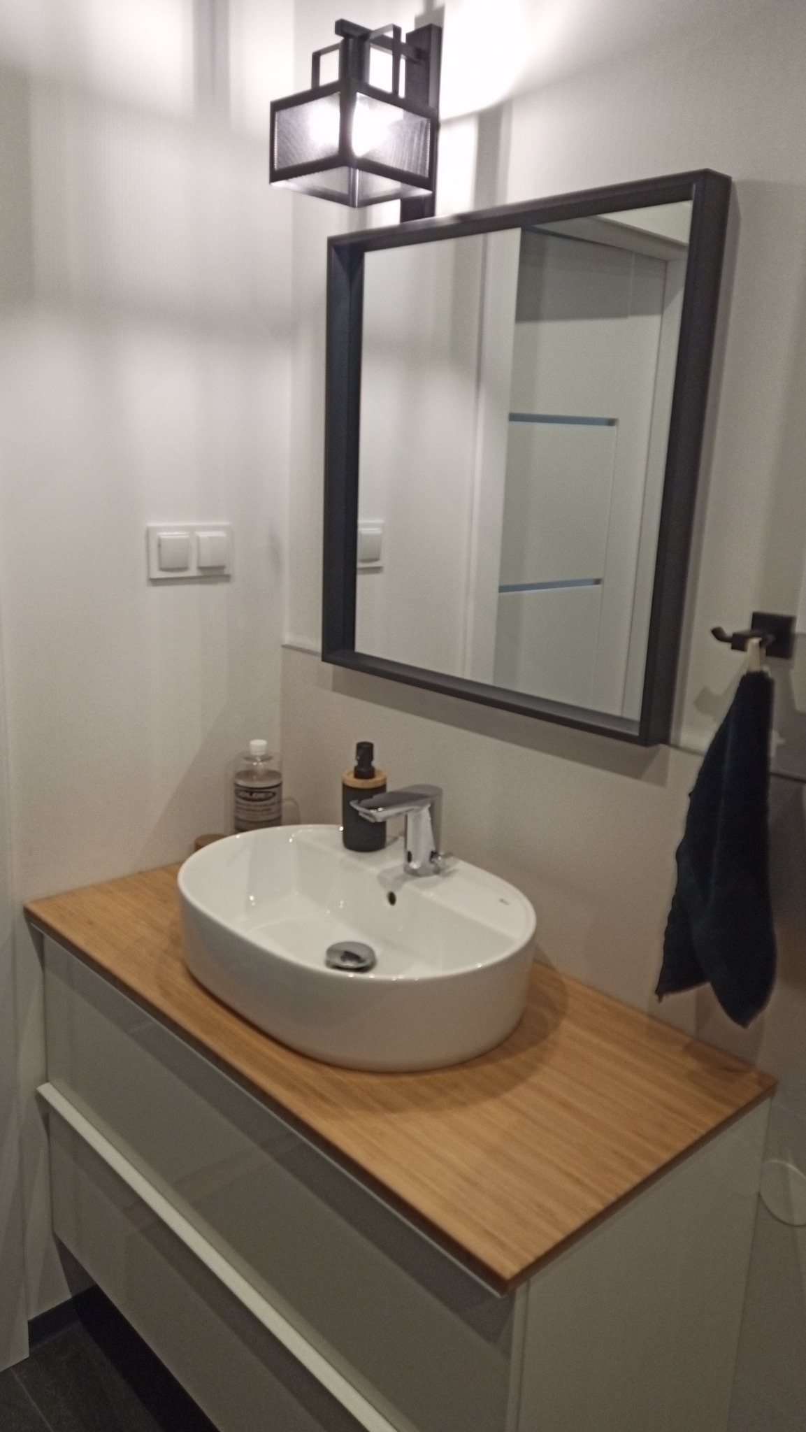 Nowoczesna łazienka z owalną umywalką nablatową, drewnianym blatem, czarnym lustrem w ramie i designerską lampą sufitową, uzupełniona o dozownik mydła i ręcznik.