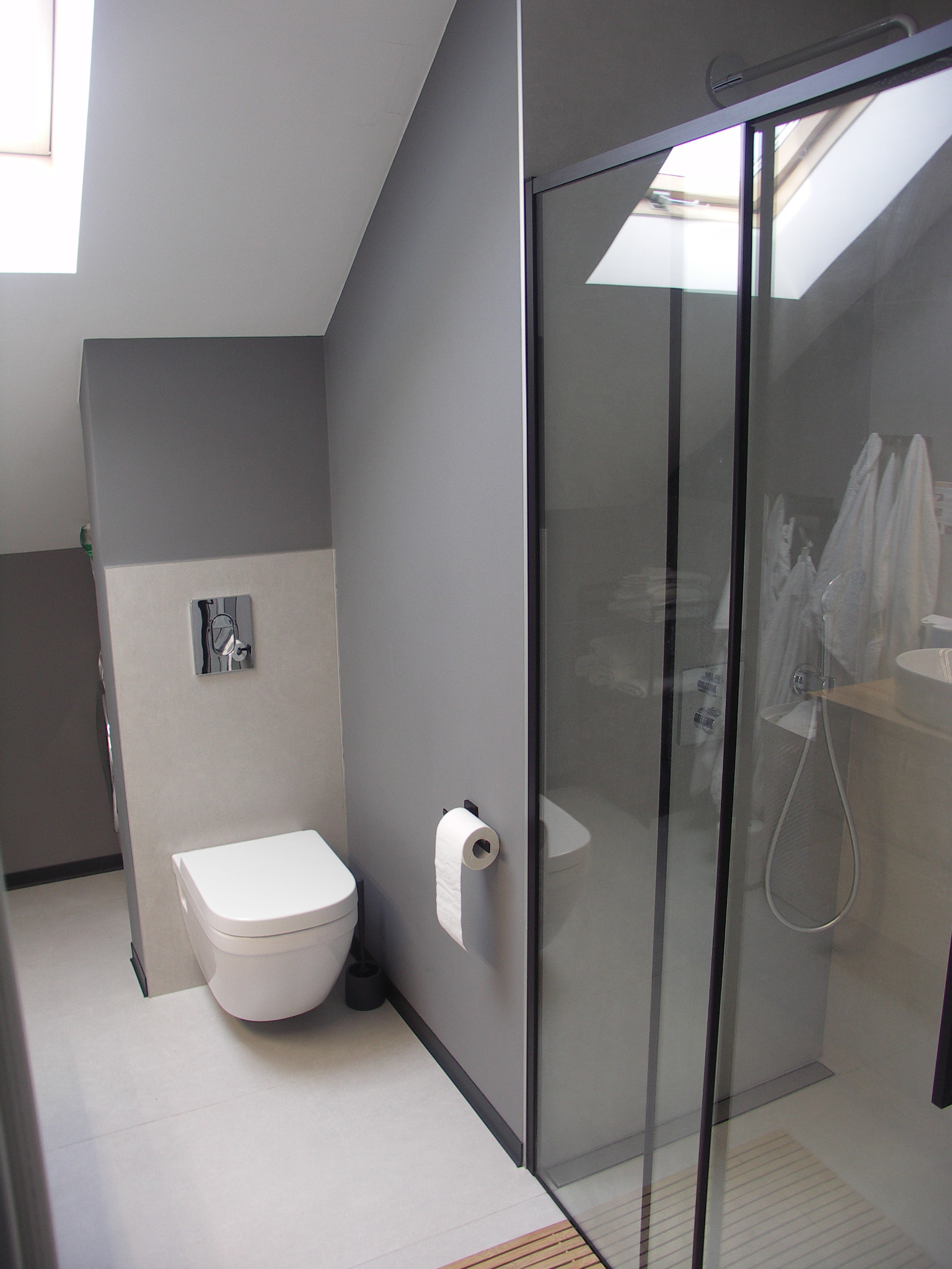 Nowoczesna łazienka na poddaszu z kabiną prysznicową typu walk-in, podwieszaną toaletą i szarą kolorystyką ścian oraz sufitowym oknem doświetlającym wnętrze.