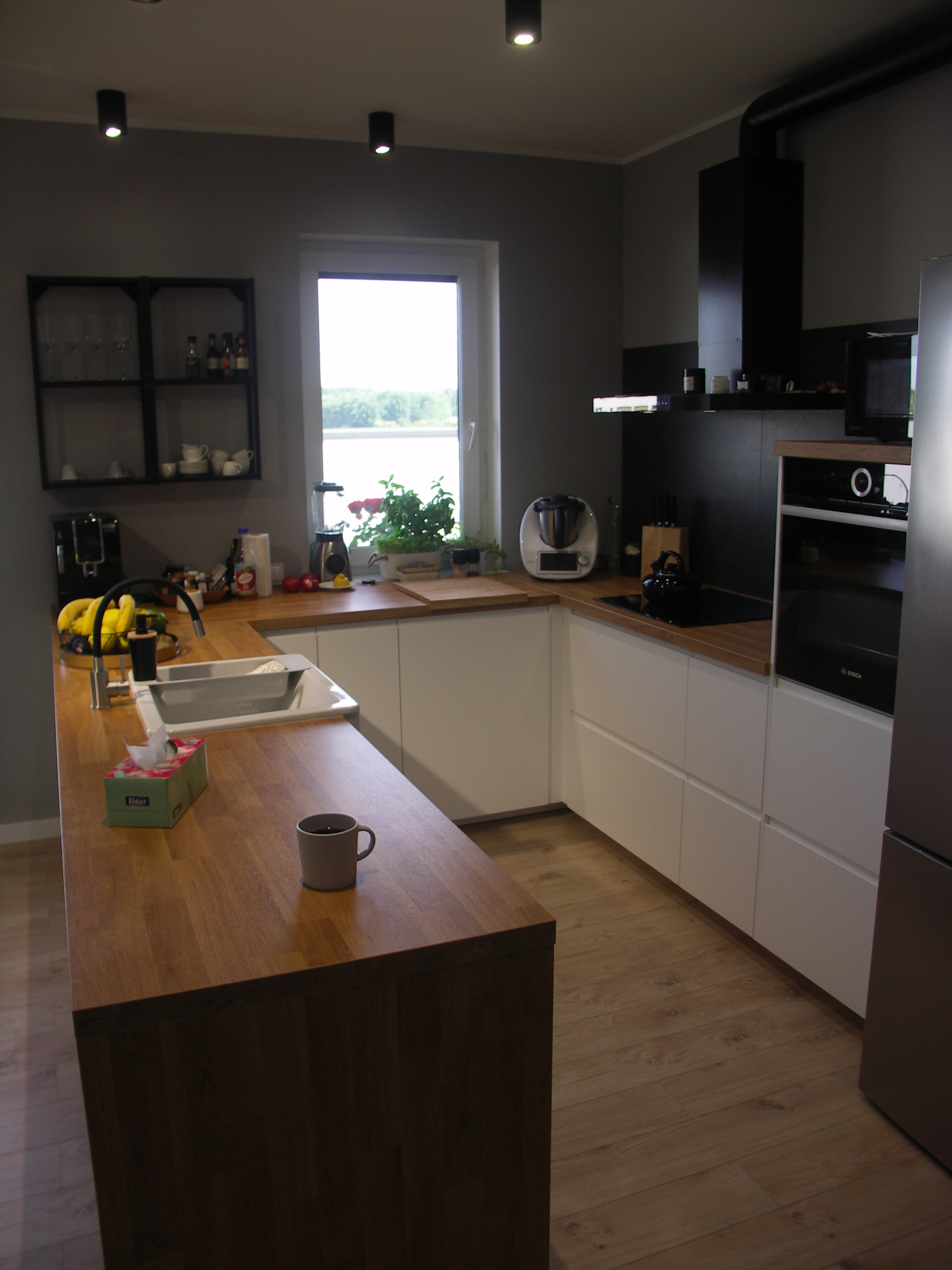 Nowoczesna kuchnia w odcieniach szarości i bieli z drewnianymi blatami, widok na okno z zielenią, na blacie stoi kubek i pudełko chusteczek.