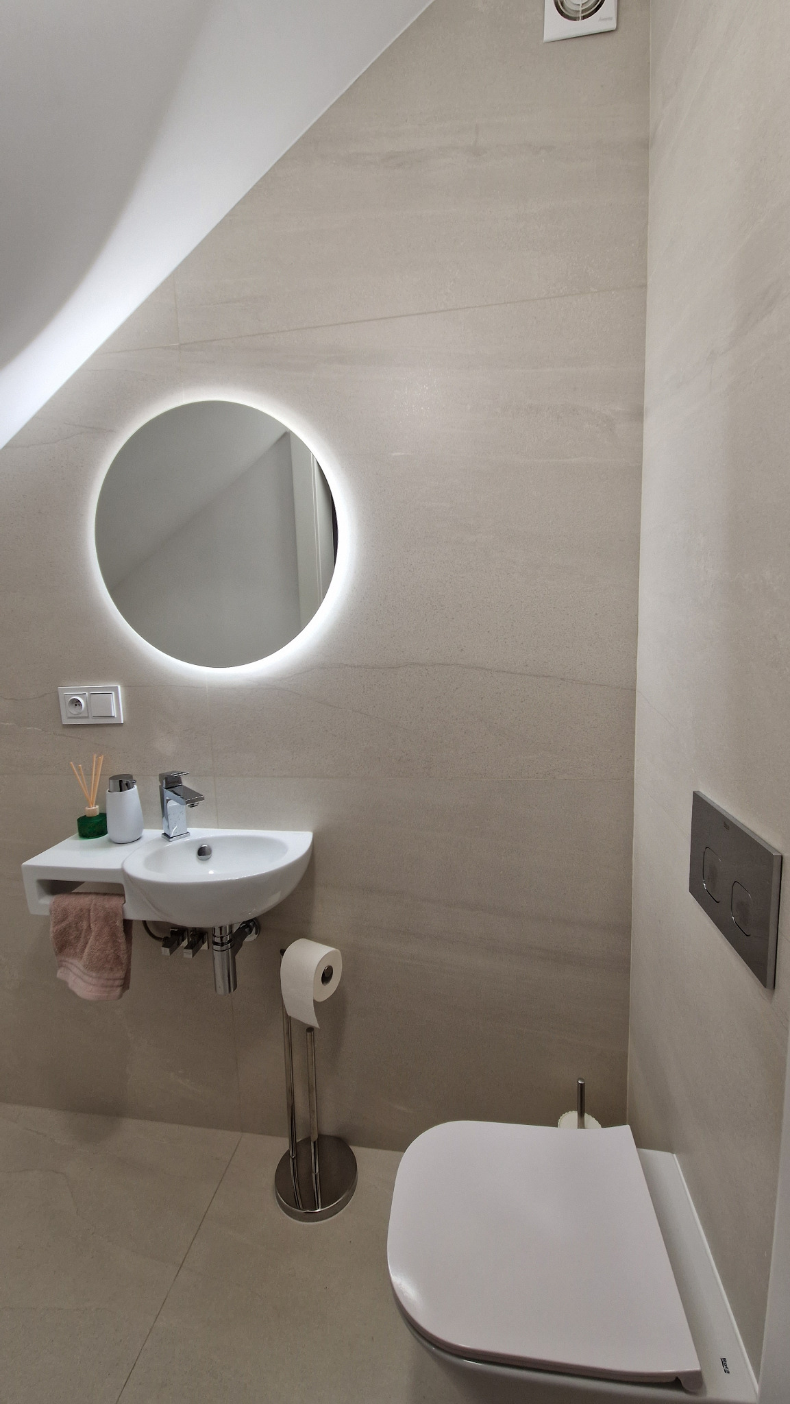 Jasna łazienka na poddaszu z beżowymi płytkami, okrągłym lustrem podświetlanym LED, wiszącą umywalką z małym blatem, toaletą i akcesoriami.
