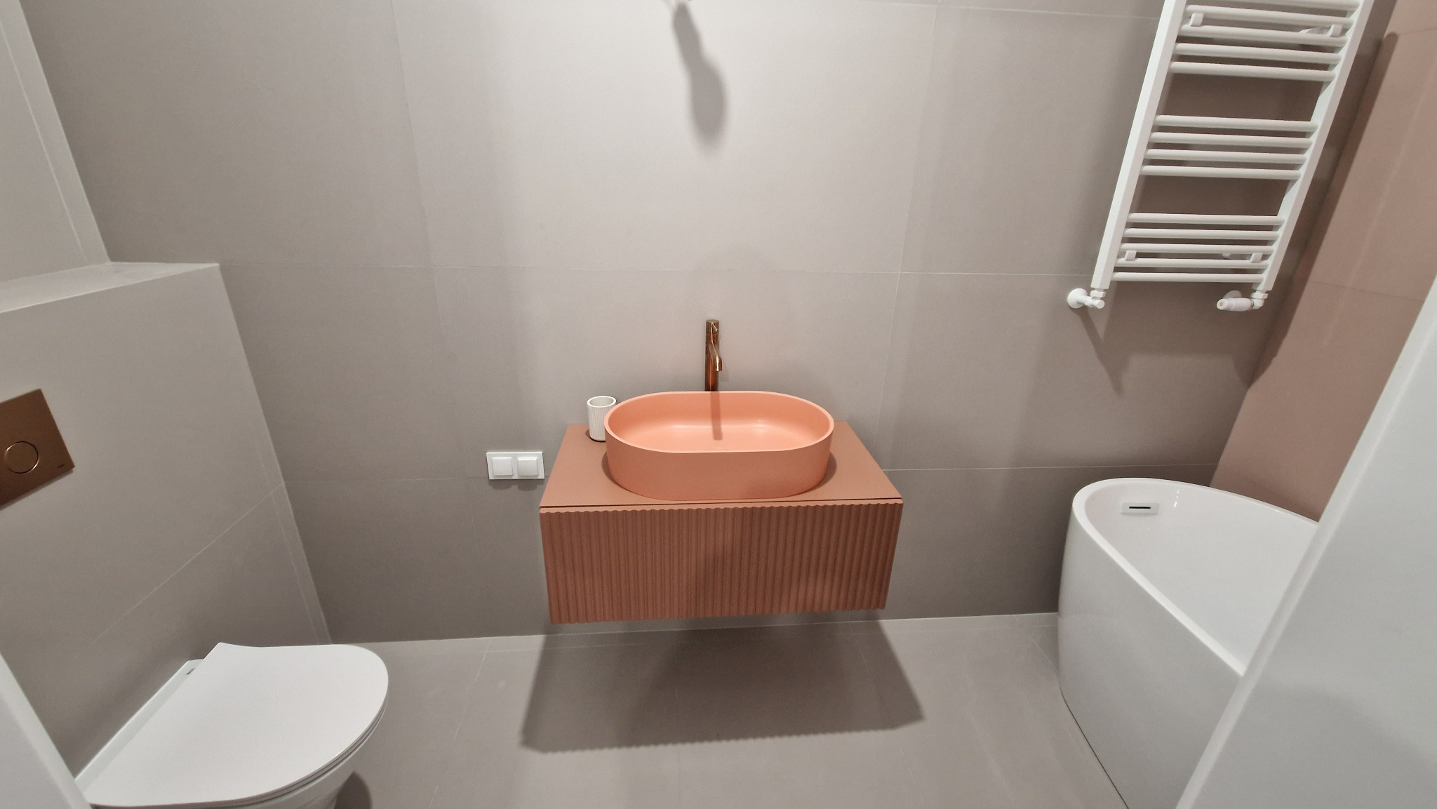 Nowoczesna łazienka z owalną, brzoskwiniową umywalką na szafce z ryflowanym frontem, złotą baterią, grzejnikiem drabinkowym i minimalistyczną toaletą podtynkową.