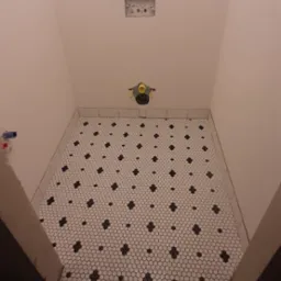 Ułożona mozaika podłogowa w łazience, białe sześciokąty z czarnymi akcentami, widoczne przygotowane przyłącza wodno-kanalizacyjne, oraz akcesoria glazurnicze na podłodze.