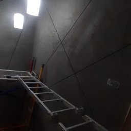 Jakub Kościelniak - Ściana pokryta szarym tynkiem dekoracyjnym z wyżłobionymi wzorami geometrycznymi, oświetlona dwoma wiszącymi lampami, z widoczną drabiną i fragmentem schodów w trakcie prac wykończeniowych.