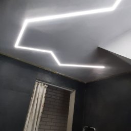 Jakub Kościelniak - Nowoczesne oświetlenie sufitowe LED w kształcie zygzaka w pokoju z ciemnymi ścianami, widoczna drabina i fragment zasłoniętego wejścia do pomieszczenia z ceglaną ścianą.