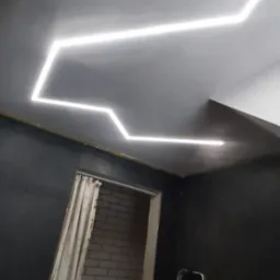 Nowoczesne oświetlenie sufitowe LED w kształcie zygzaka w pokoju z ciemnymi ścianami, widoczna drabina i fragment zasłoniętego wejścia do pomieszczenia z ceglaną ścianą.