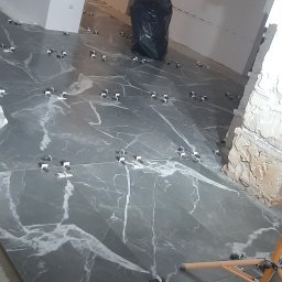 Jakub Kościelniak - Układanie płytek imitujących marmur na podłodze w trakcie remontu, widoczne klipsy poziomujące i fragment statywu, w tle czarny worek na śmieci i fragment ściany z niedokończonym wykończeniem.