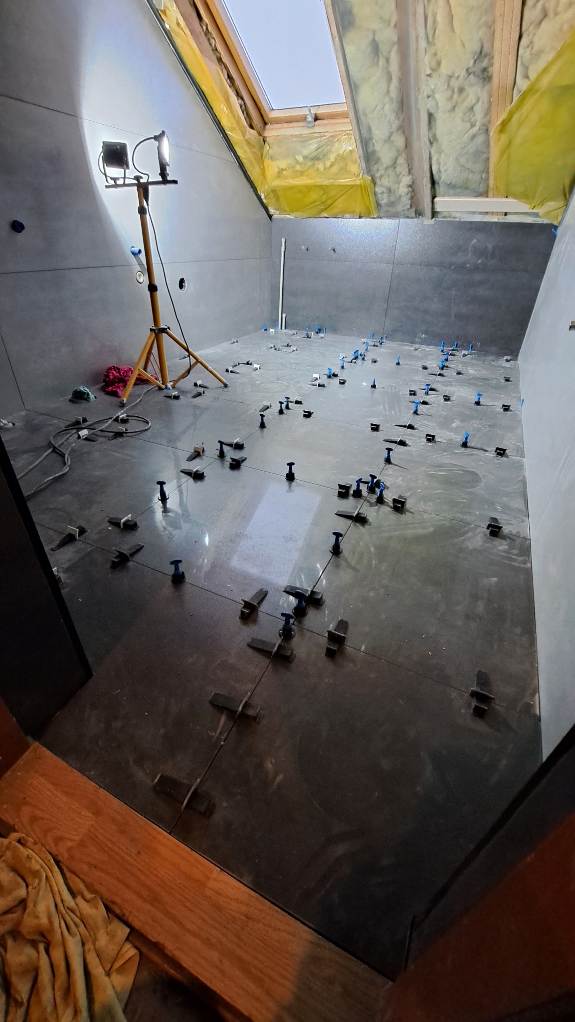 Montaż ciemnych płytek podłogowych w łazience na poddaszu ze świetlikiem; widoczne narzędzia do poziomowania i lampa budowlana.