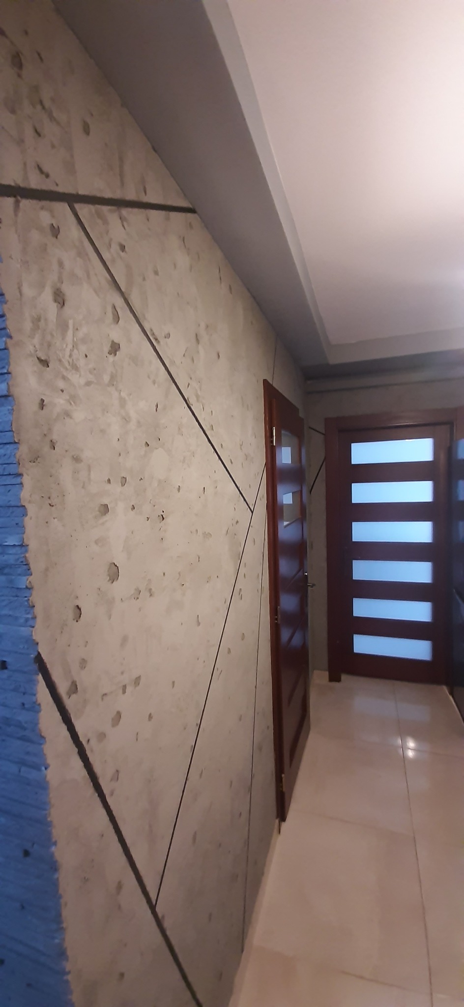 Ściana wewnętrzna z dekoracyjnym tynkiem imitującym beton, z widocznymi liniami podziału i fragmentem ściany z niebieskim materiałem izolacyjnym, w tle widoczne drzwi wewnętrzne z przeszkleniami.