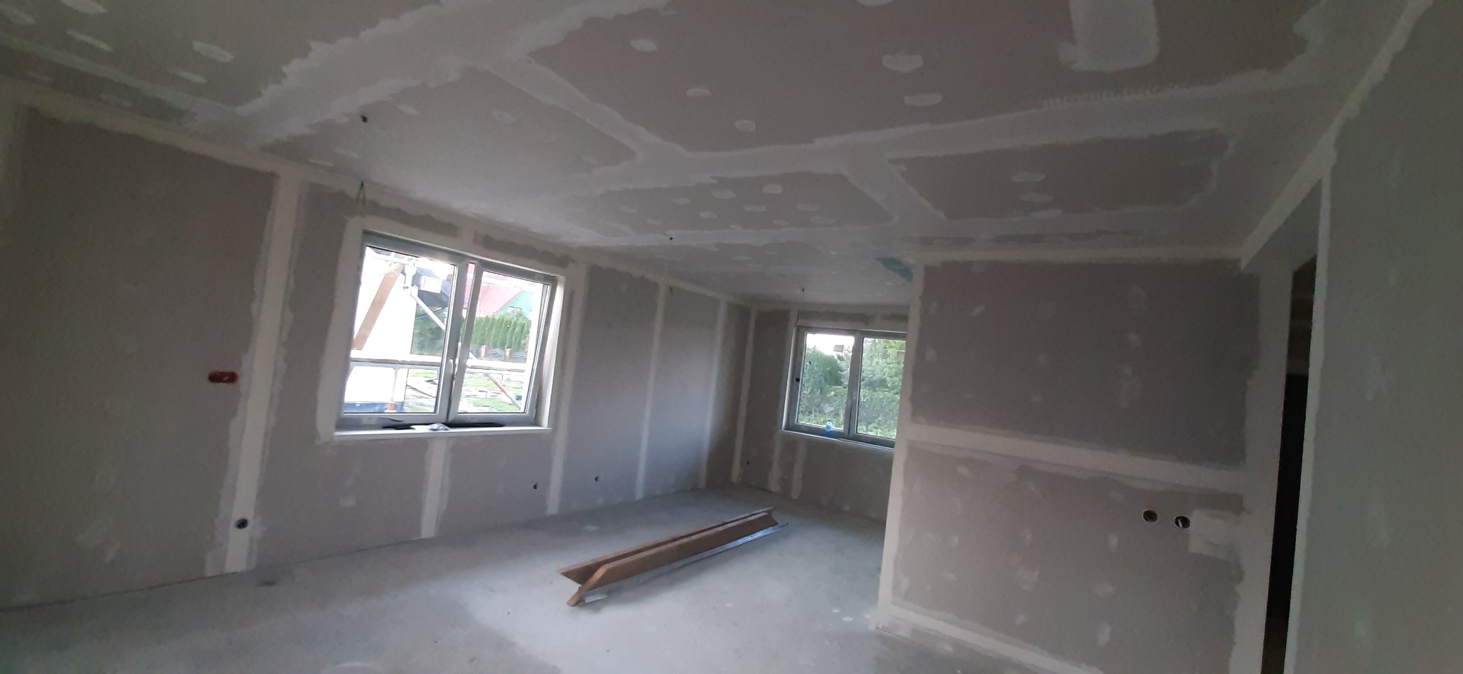 Wnętrze w trakcie prac wykończeniowych: ściany i sufit pokryte płytami kartonowo-gipsowymi, widoczne łączenia szpachlowane, dwa okna z widokiem na zieleń na zewnątrz, na podłodze leżą profile...