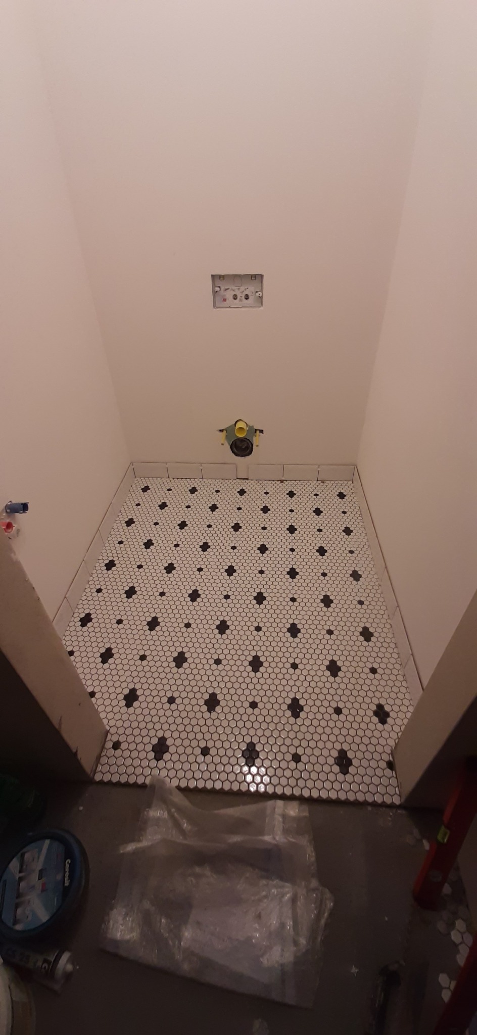 Ułożona mozaika podłogowa w łazience, białe sześciokąty z czarnymi akcentami, widoczne przygotowane przyłącza wodno-kanalizacyjne, oraz akcesoria glazurnicze na podłodze.