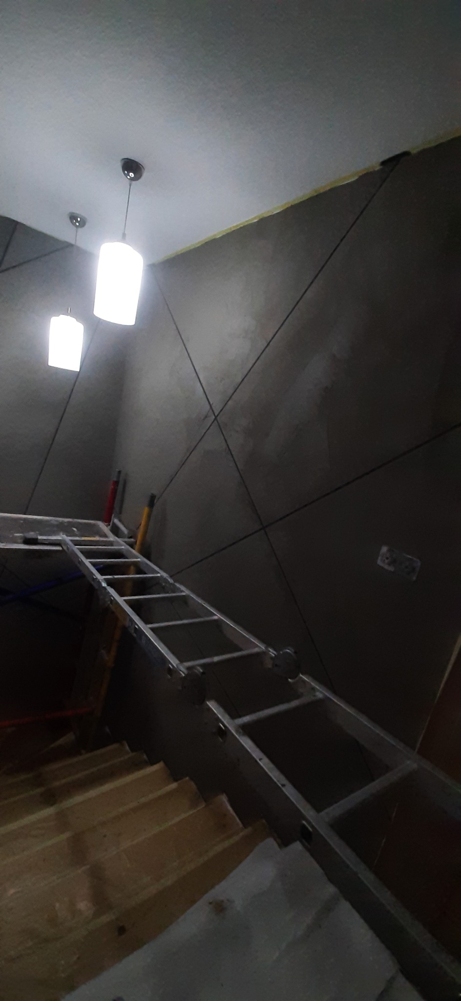 Ściana pokryta szarym tynkiem dekoracyjnym z wyżłobionymi wzorami geometrycznymi, oświetlona dwoma wiszącymi lampami, z widoczną drabiną i fragmentem schodów w trakcie prac wykończeniowych.