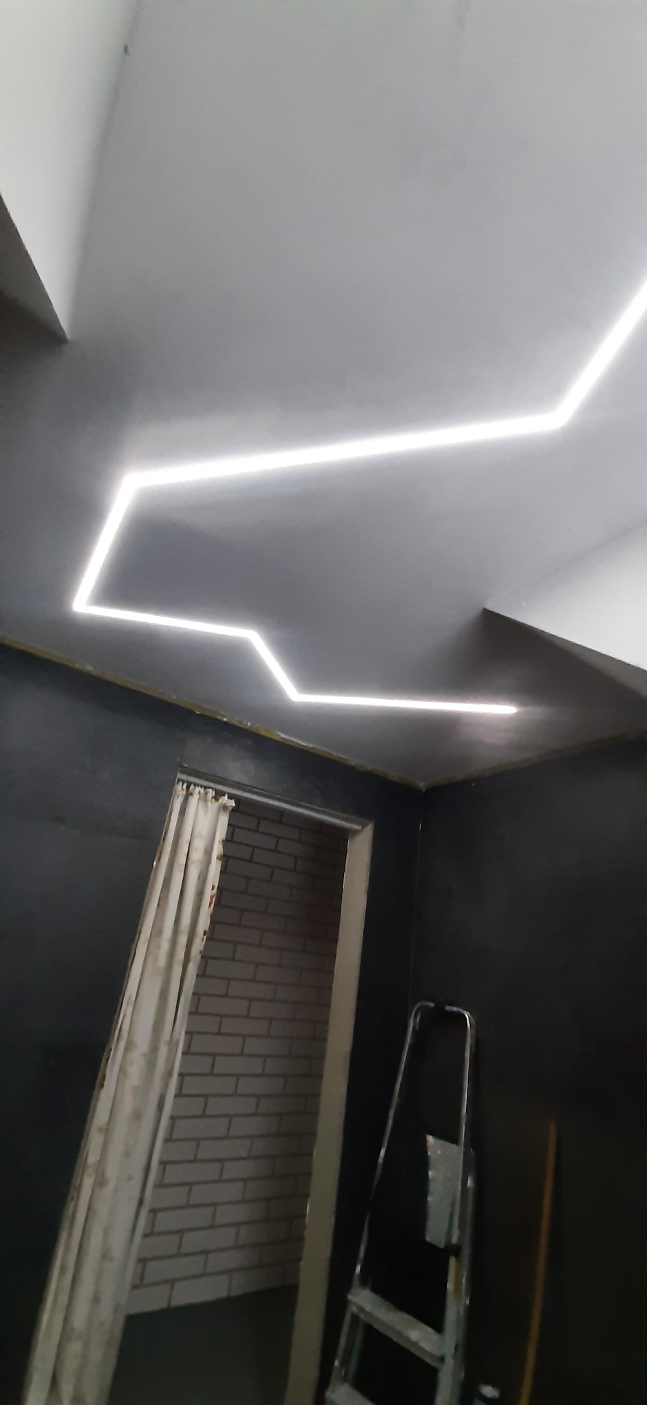 Nowoczesne oświetlenie sufitowe LED w kształcie zygzaka w pokoju z ciemnymi ścianami, widoczna drabina i fragment zasłoniętego wejścia do pomieszczenia z ceglaną ścianą.