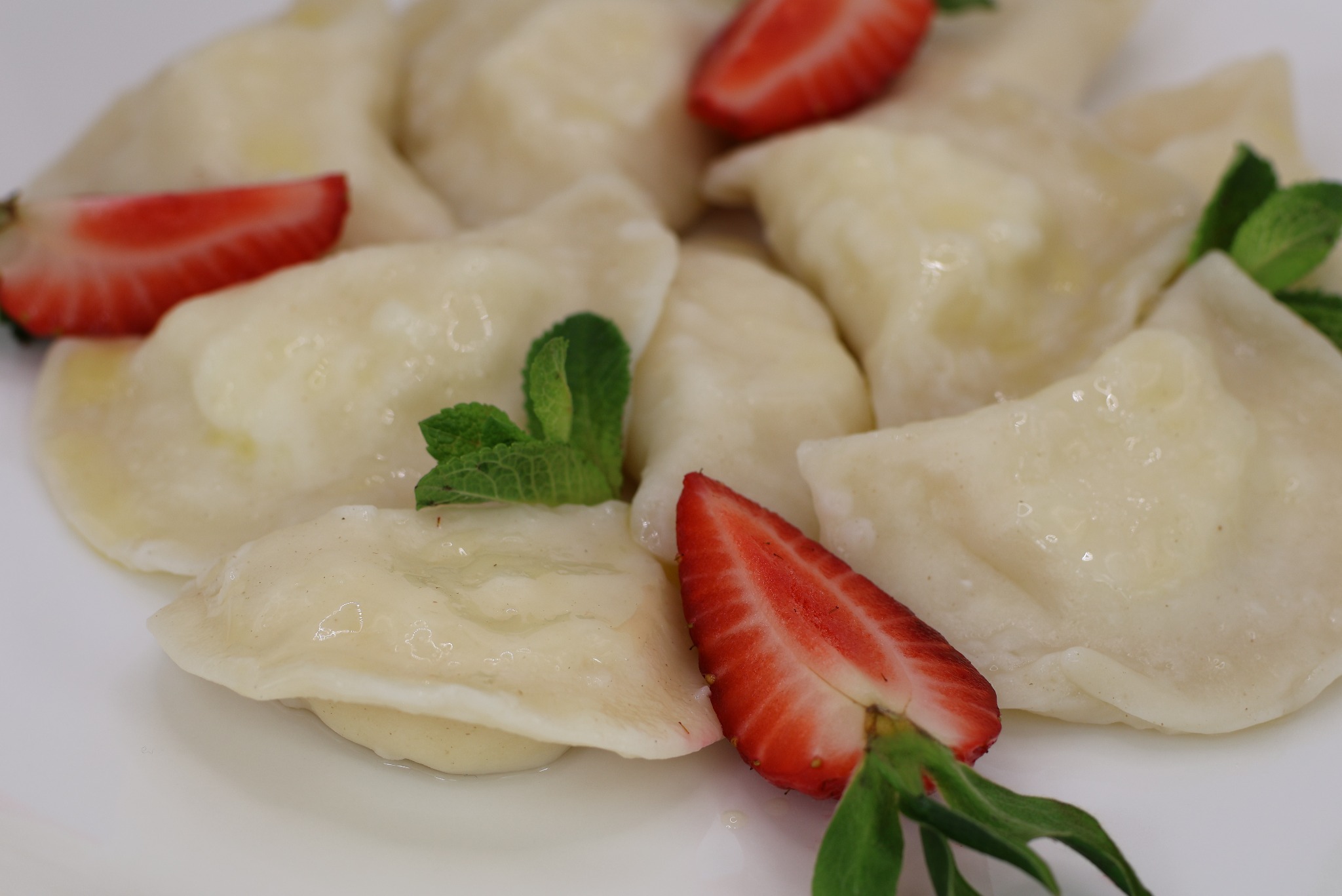 Delikatne pierogi polane masłem, udekorowane świeżymi truskawkami w plasterkach i listkami mięty na białym talerzu.