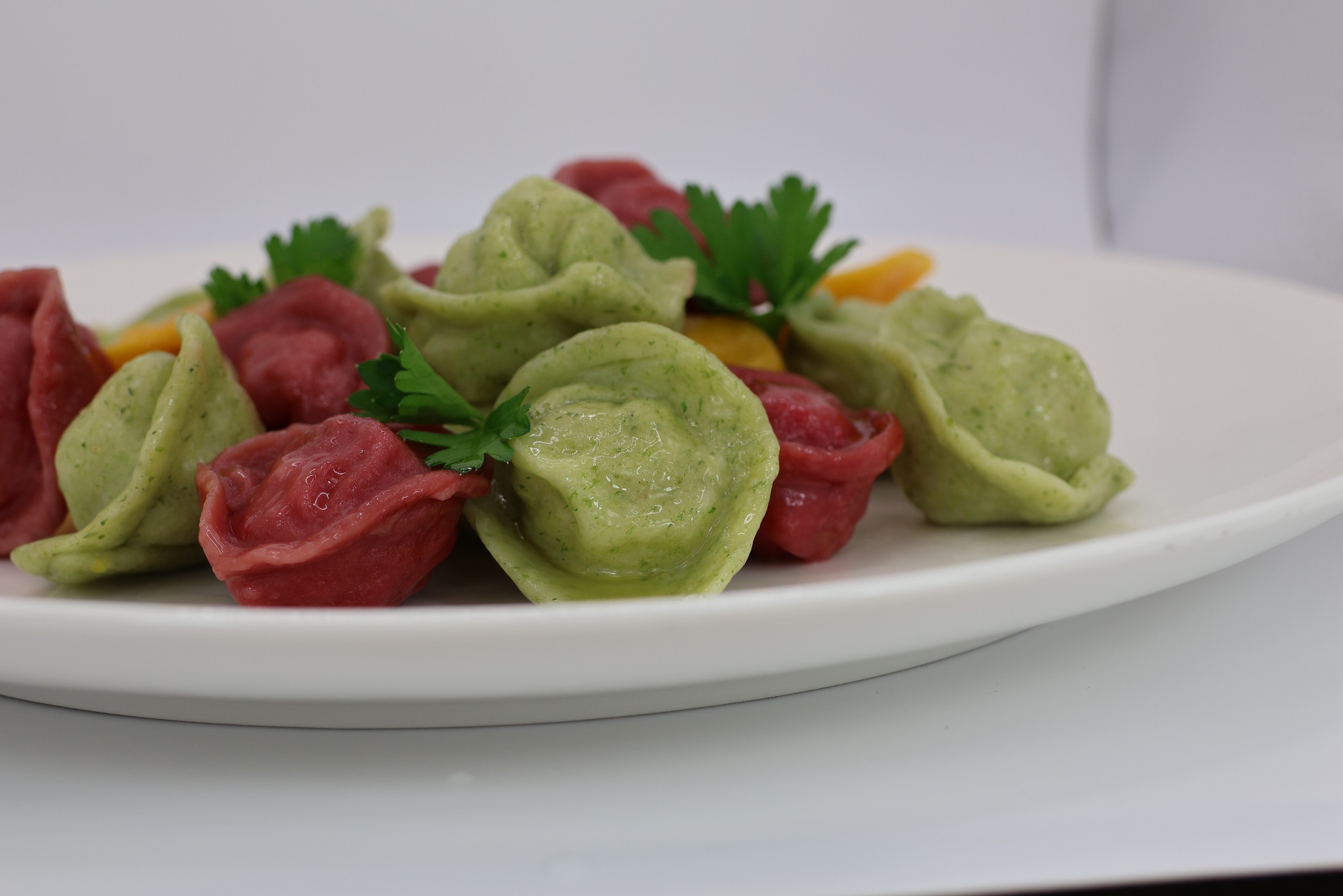 Ręcznie robione, barwione naturalnie pierożki o kształcie sakiewki w kolorach zielonym i czerwonym, ułożone na białym talerzu, udekorowane świeżą natką pietruszki.