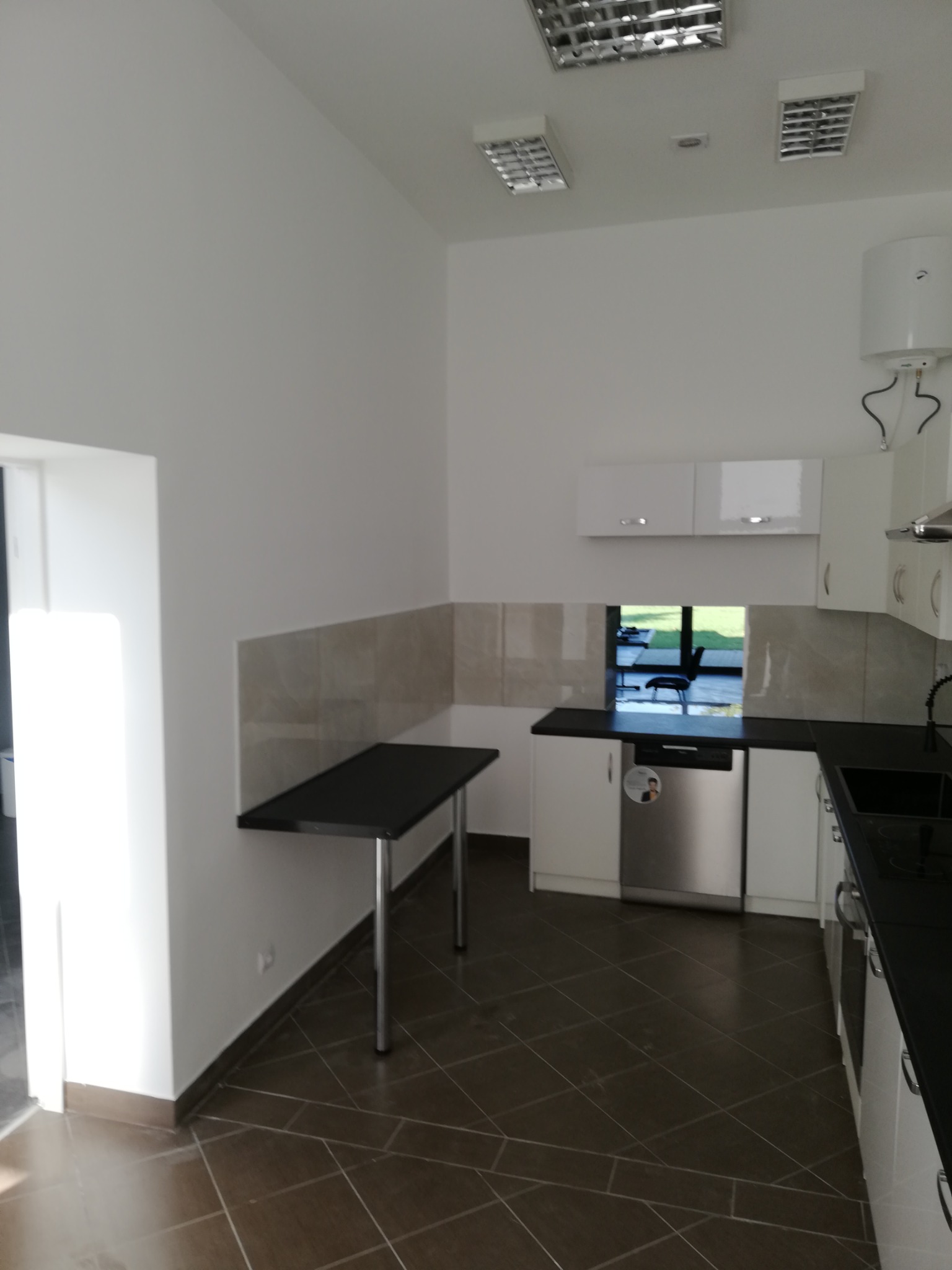 Nowoczesna, jasna kuchnia z białymi szafkami, czarnym blatem i brązową podłogą z płytek. Widoczny fragment stołu z czarnym blatem i metalowymi nogami. Okno w tle z widokiem na zieleń.