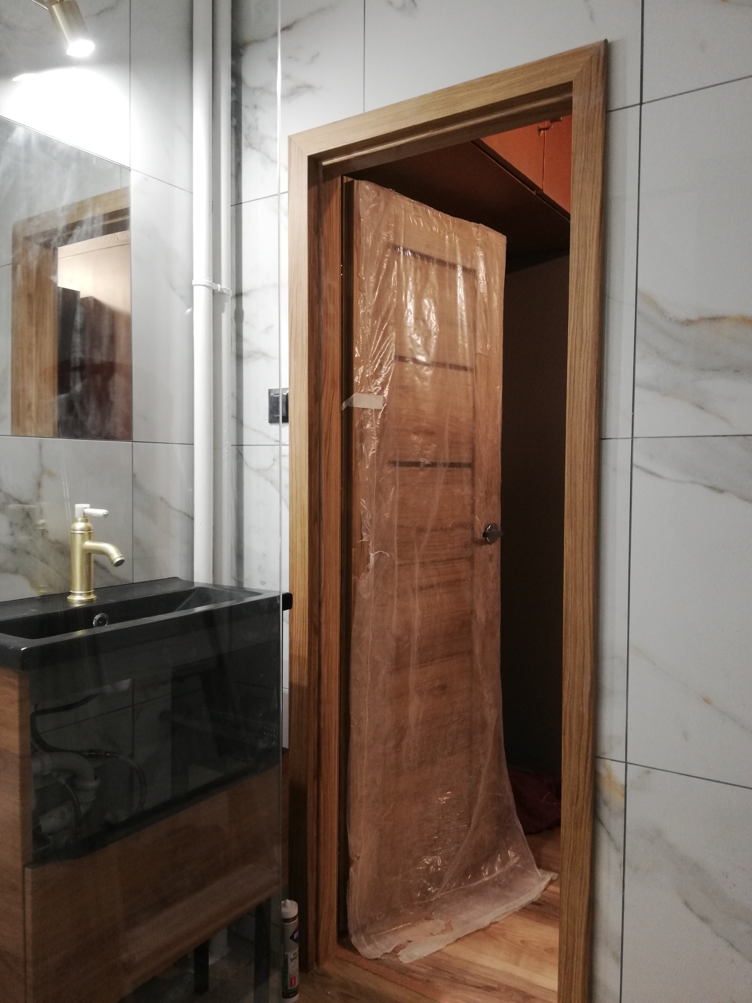 Nowa, drewniana ościeżnica z drzwiami w folii ochronnej, zamontowana w łazience z marmurowymi płytkami i czarną umywalką z złotą baterią.