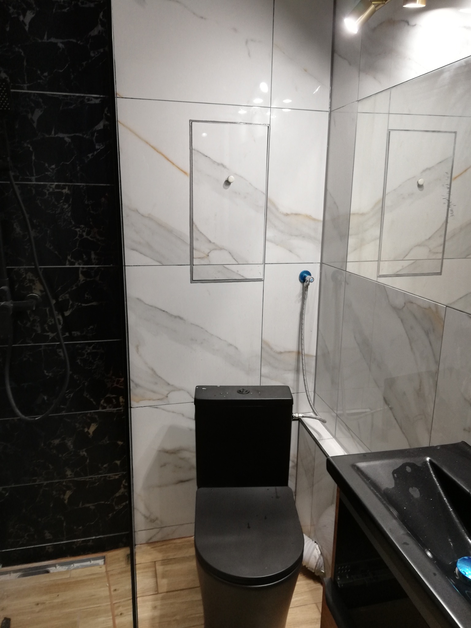 Nowoczesna łazienka z czarną armaturą: wisząca toaleta i umywalka. Ściany wyłożone białymi płytkami imitującymi marmur z elementami czarnego marmuru. Ukryta rewizja za toaletą.