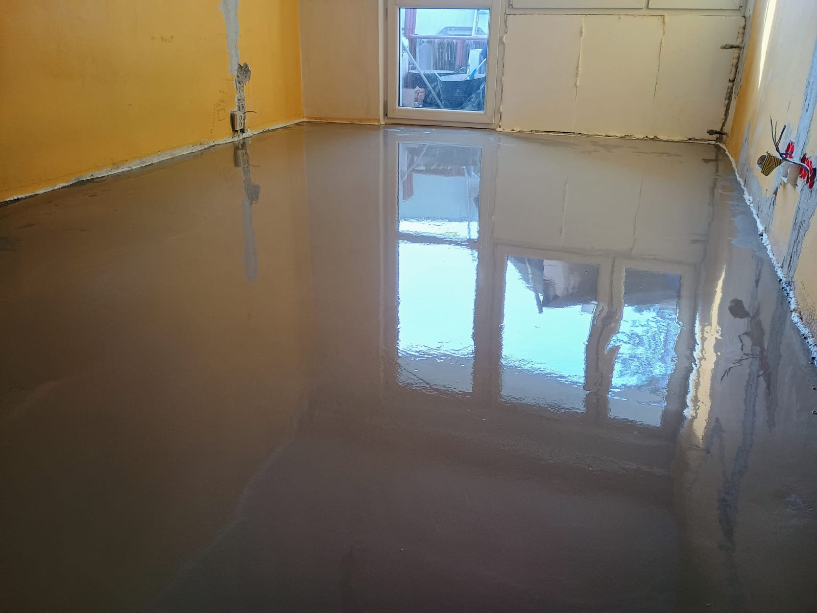 Świeżo wylana, samopoziomująca wylewka podłogowa w pomieszczeniu z żółtymi i białymi ścianami, widoczne instalacje elektryczne.