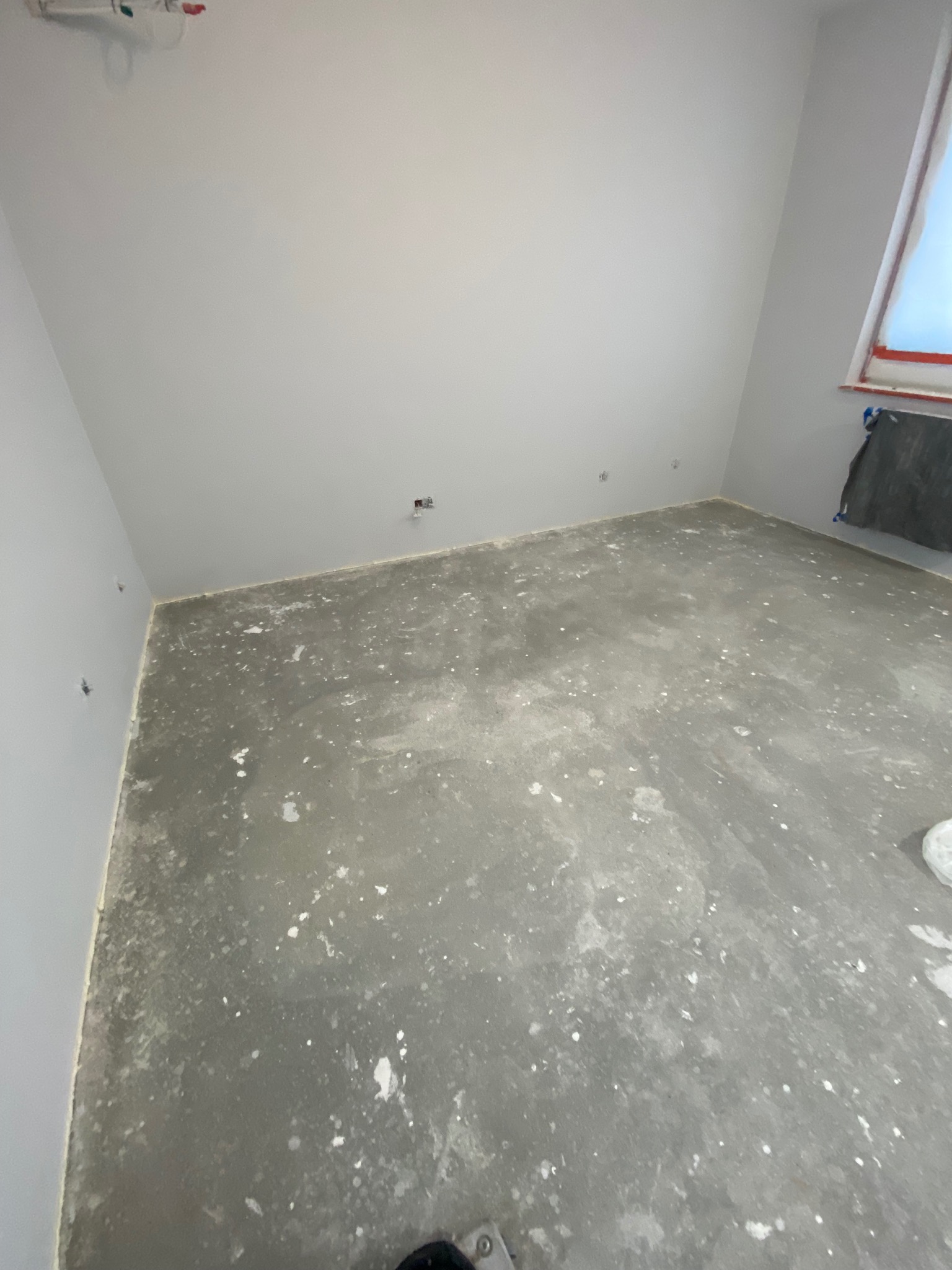 Pokój w trakcie remontu: szare ściany pomalowane, surowa betonowa podłoga z plamami farby, widoczne puszki elektryczne w ścianie, okno zabezpieczone folią.