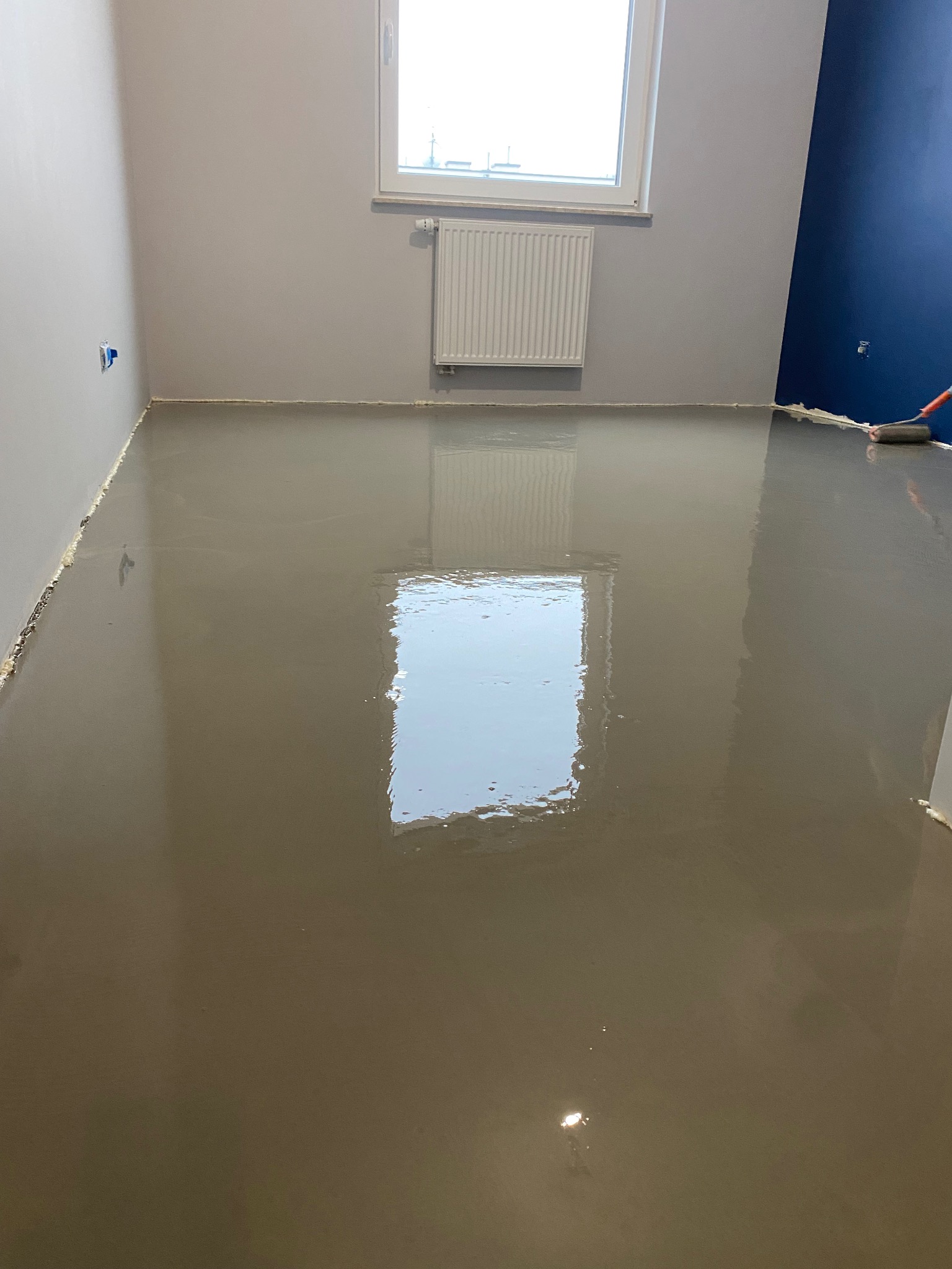 Wylewka samopoziomująca w trakcie aplikacji, widoczny wałek malarski i świeżo wylana, gładka powierzchnia podłogi w szarym pomieszczeniu z oknem i grzejnikiem.