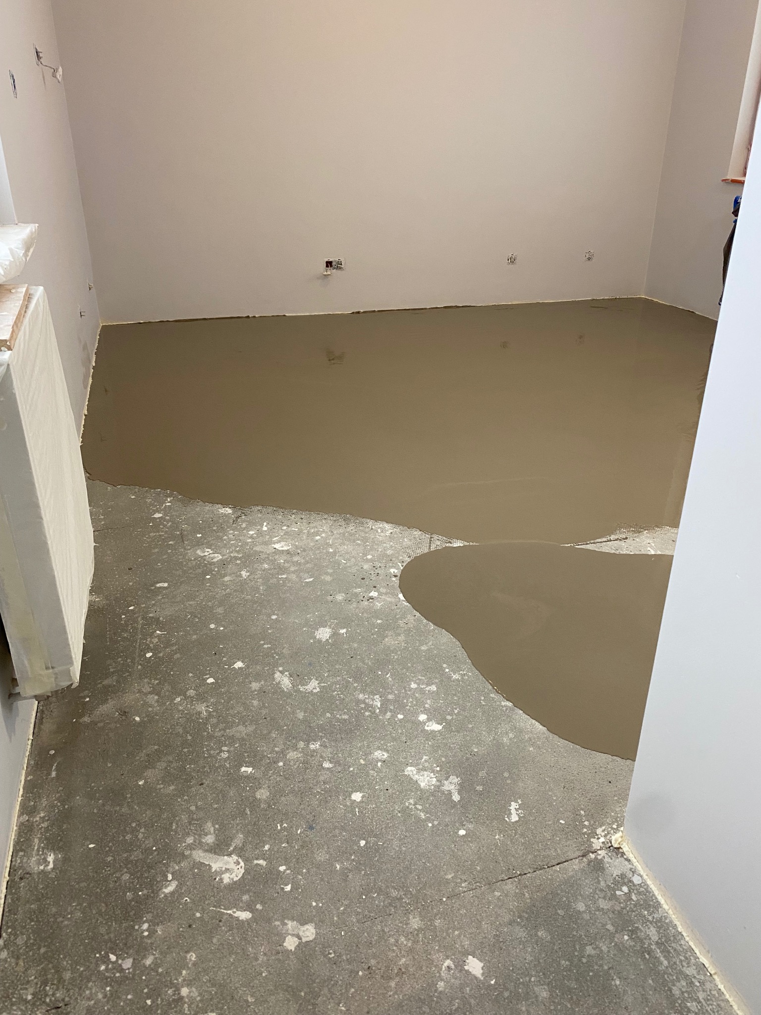 Wylewka samopoziomująca częściowo rozprowadzona na betonowej podłodze w pomieszczeniu z białymi ścianami i widocznymi puszkami elektrycznymi.