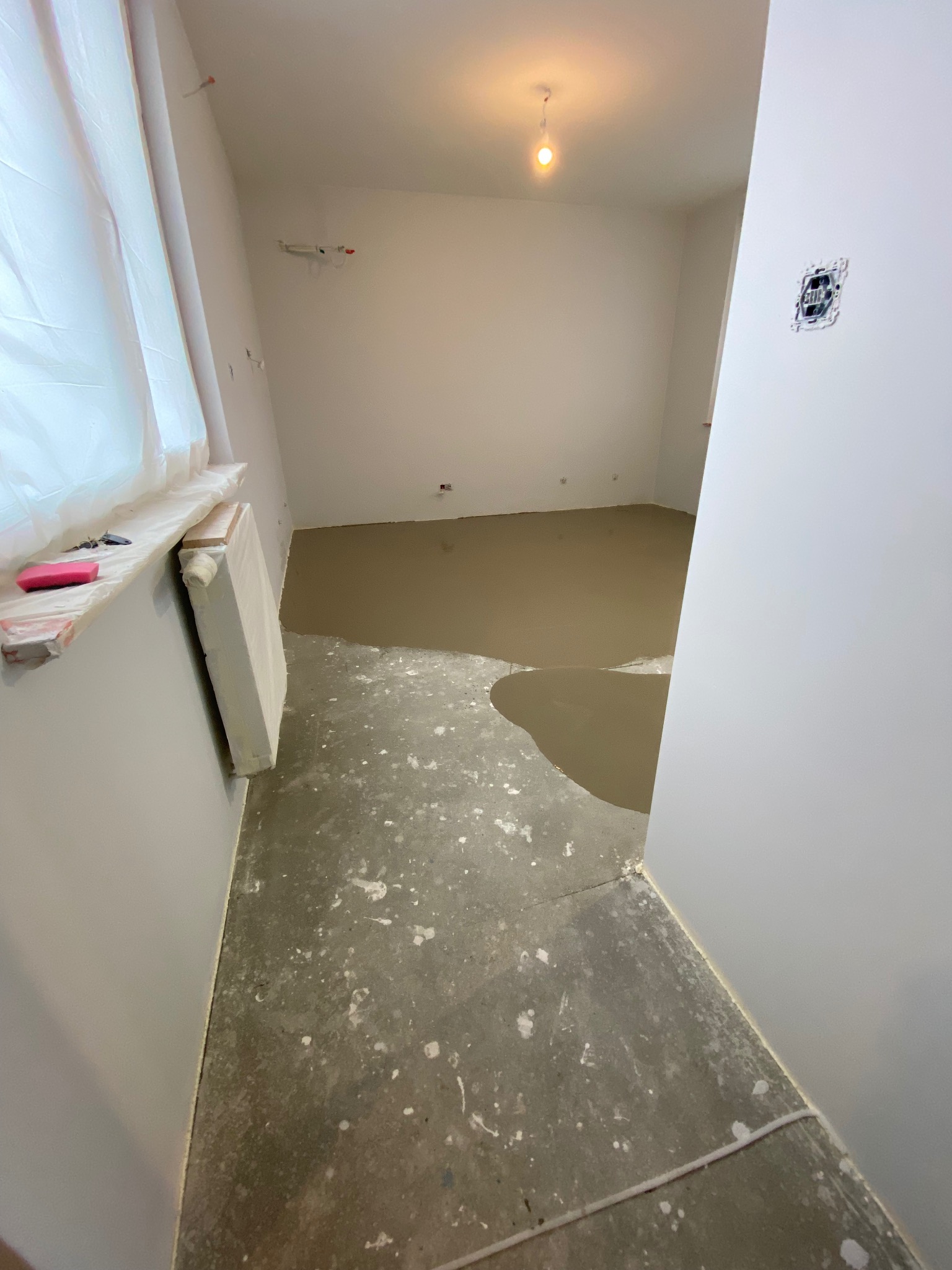 Pokój w trakcie remontu: wylany samopoziomujący podkład podłogowy, zabezpieczone okno folią, widoczna puszka elektryczna w ścianie.