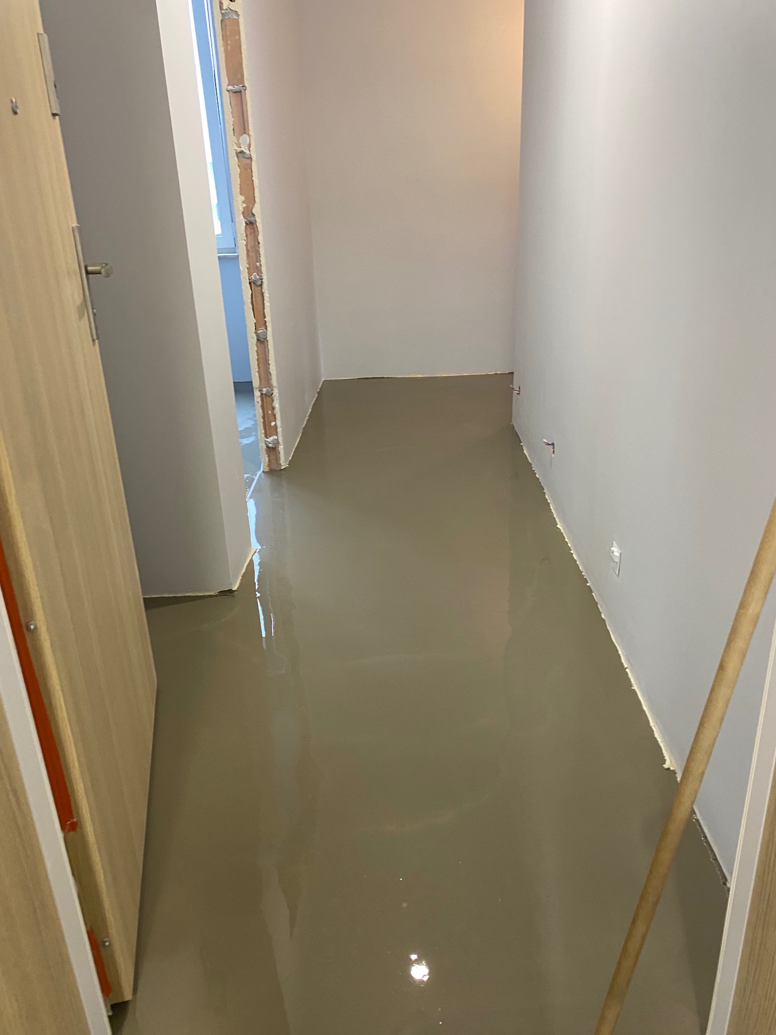 Świeżo wylana, samopoziomująca wylewka podłogowa w korytarzu podczas remontu mieszkania, widoczne ślady połatanych ścian i brak wykończenia ościeżnicy.