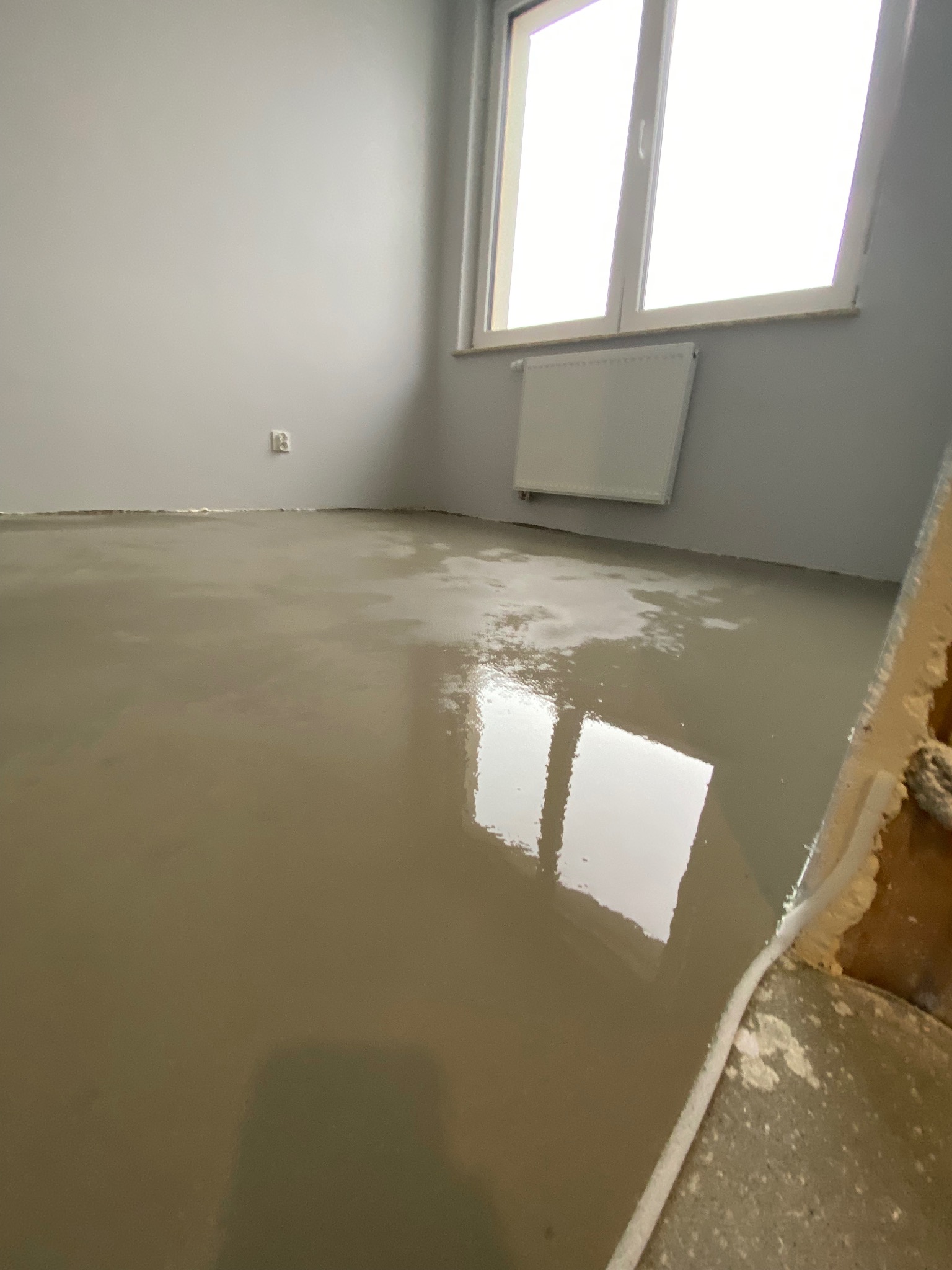 Świeżo wylana, samopoziomująca wylewka podłogowa w szarym pomieszczeniu w Krakowie, z widocznym oknem i grzejnikiem, częściowo zakryta szpachelką.