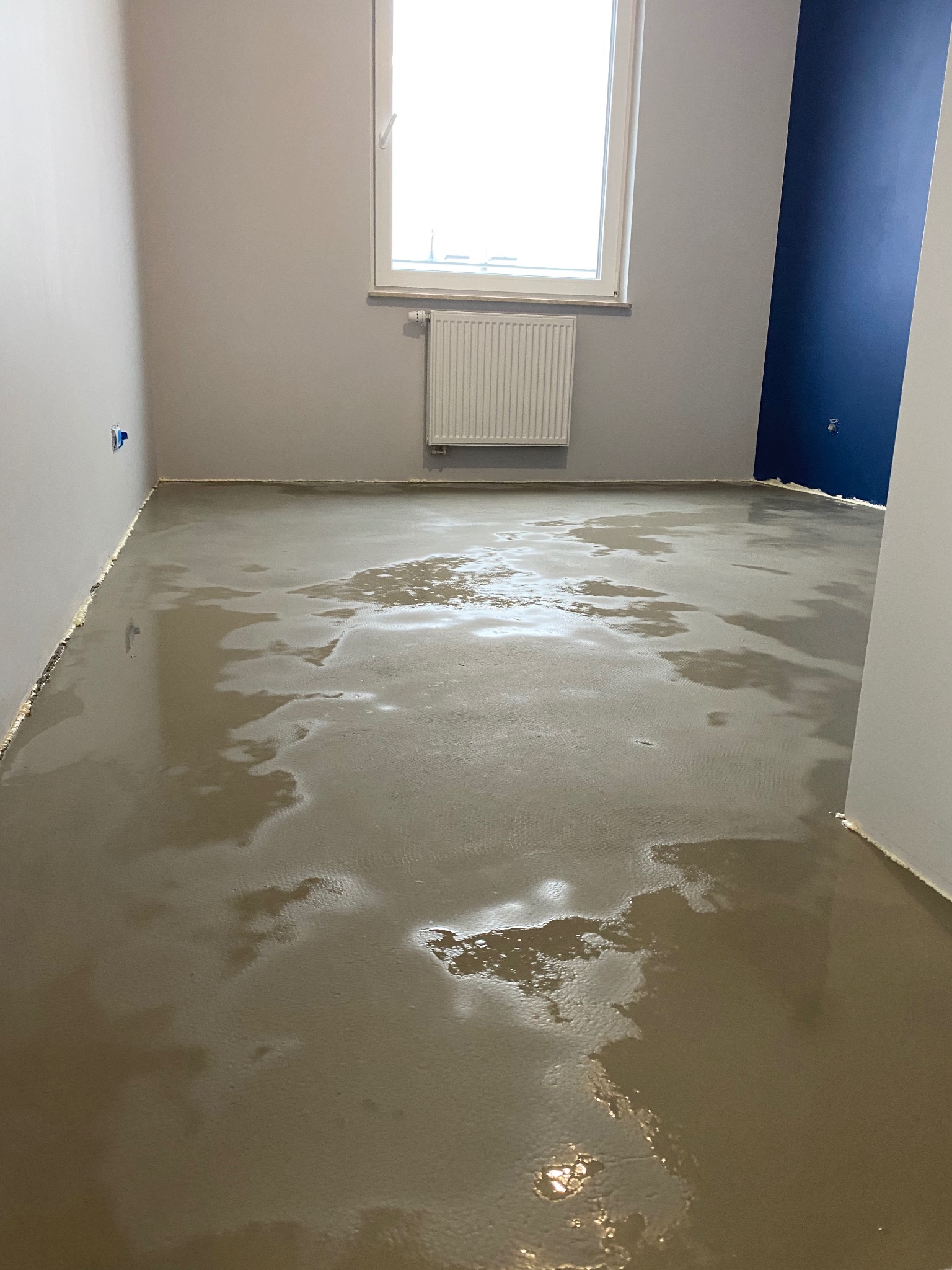 Wylewka samopoziomująca na podłodze w pomieszczeniu z grzejnikiem pod oknem i fragmentem ściany pomalowanej na granatowo.