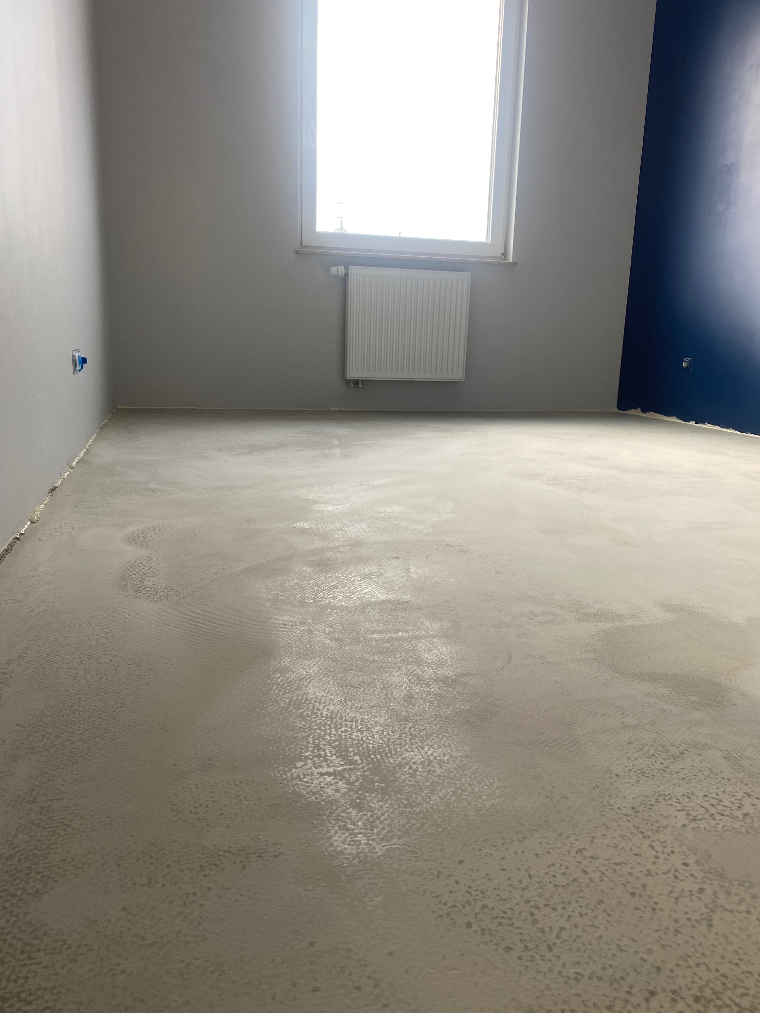 Świeżo wylana, wilgotna wylewka samopoziomująca w szarym odcieniu, z widoczną fakturą i refleksami światła, w pustym pokoju z grzejnikiem pod oknem i fragmentem ściany pomalowanej na granatowo.