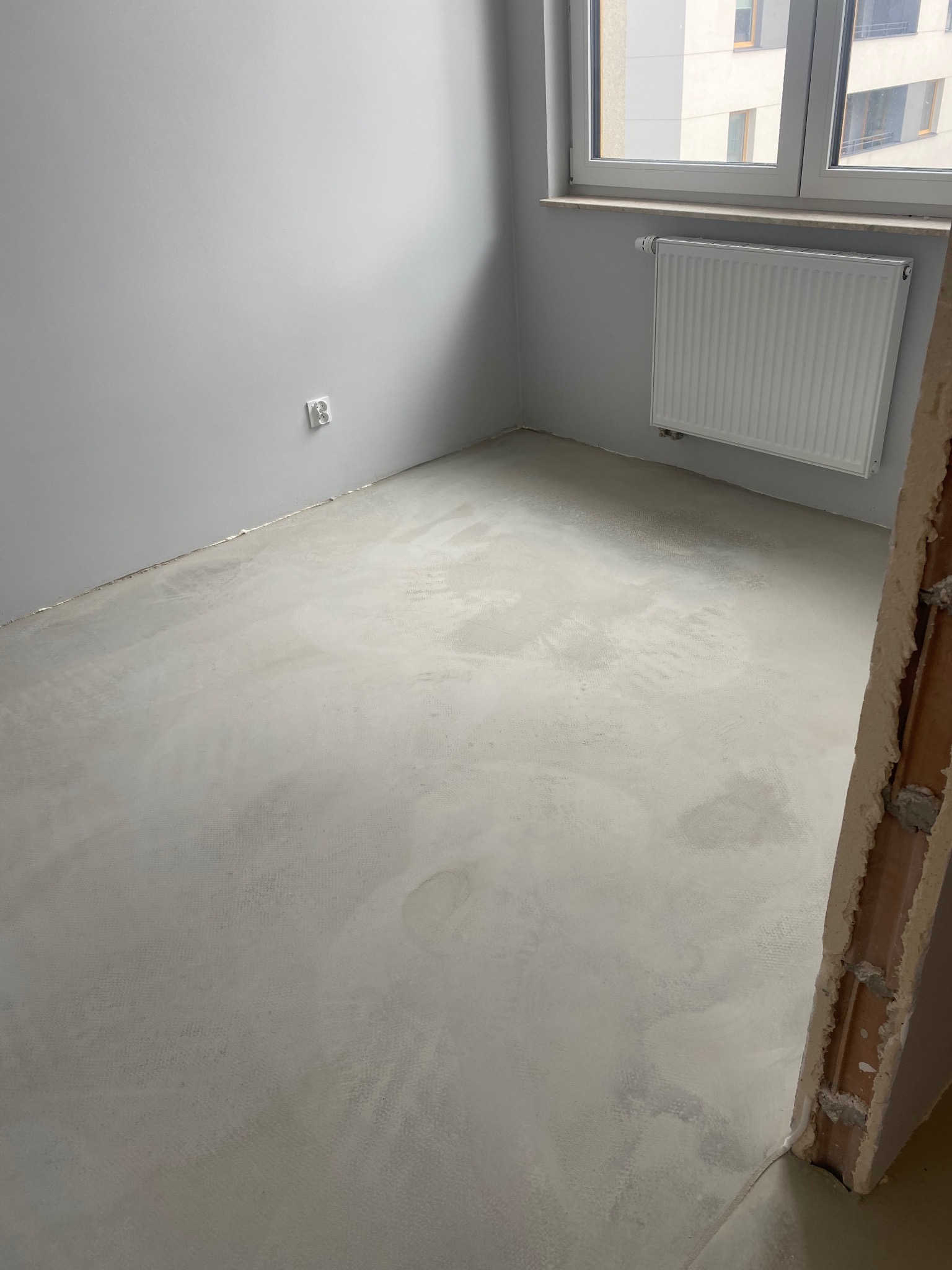 Wylewka samopoziomująca na podłodze w szarym pokoju z grzejnikiem pod oknem i gniazdkiem elektrycznym na ścianie. Widoczny fragment surowego muru w rogu.