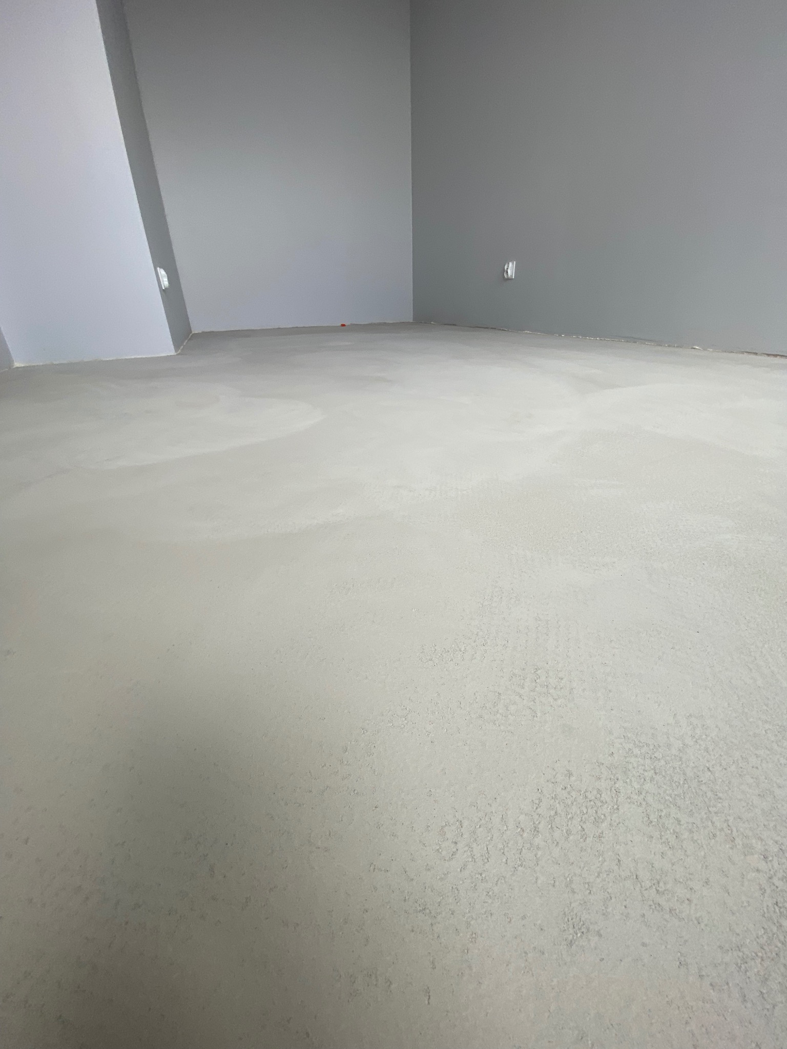 Wylany, świeży, jasnoszary betonowy podkład podłogowy w pustym, szarym pomieszczeniu z widocznymi gniazdkami elektrycznymi na ścianie.