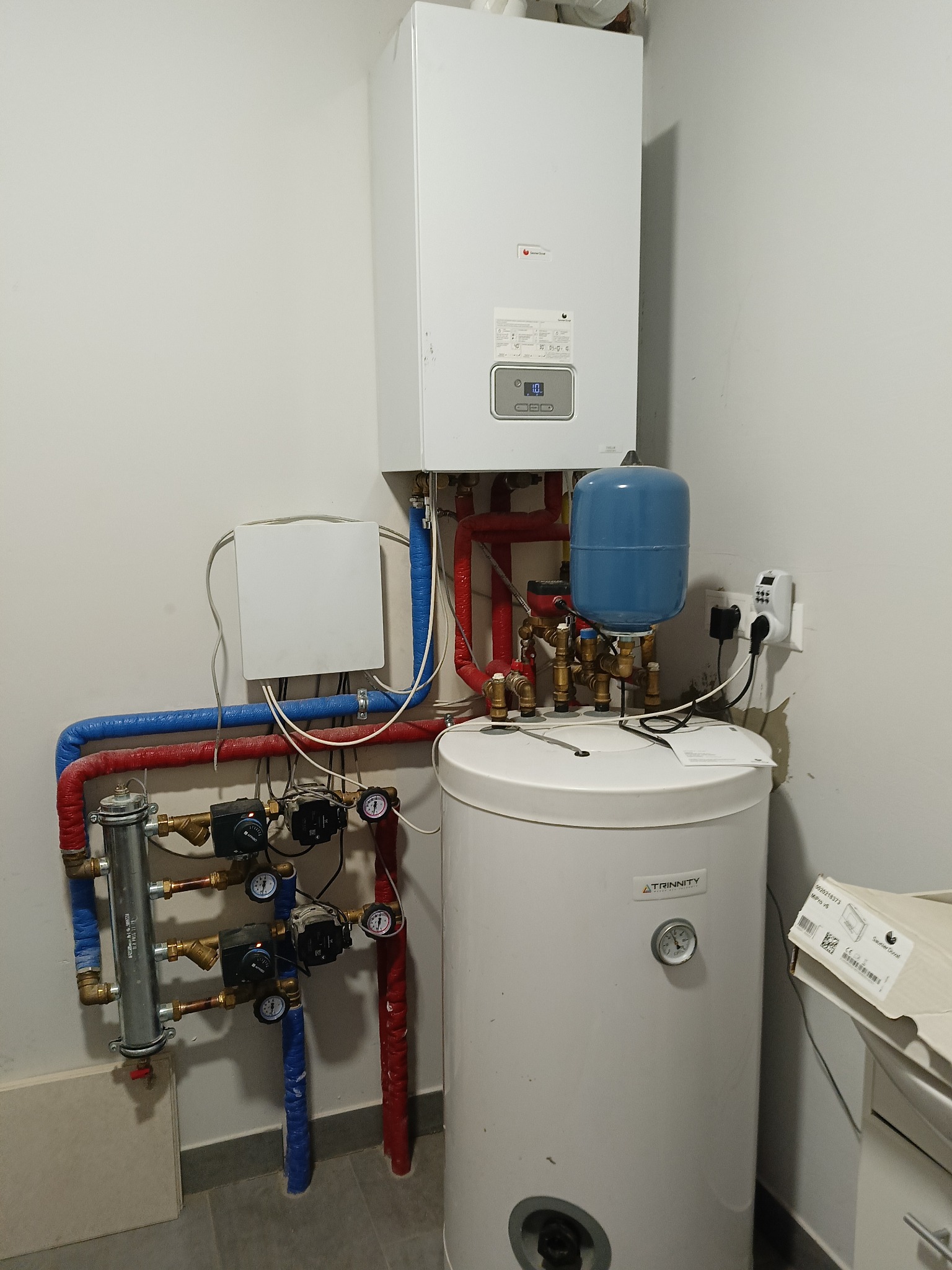 Biały piec gazowy kondensacyjny z podłączonym zasobnikiem ciepłej wody i instalacją hydrauliczną z rurami w izolacji czerwonej i niebieskiej, widoczny manometr i elektroniczny sterownik temperatury.