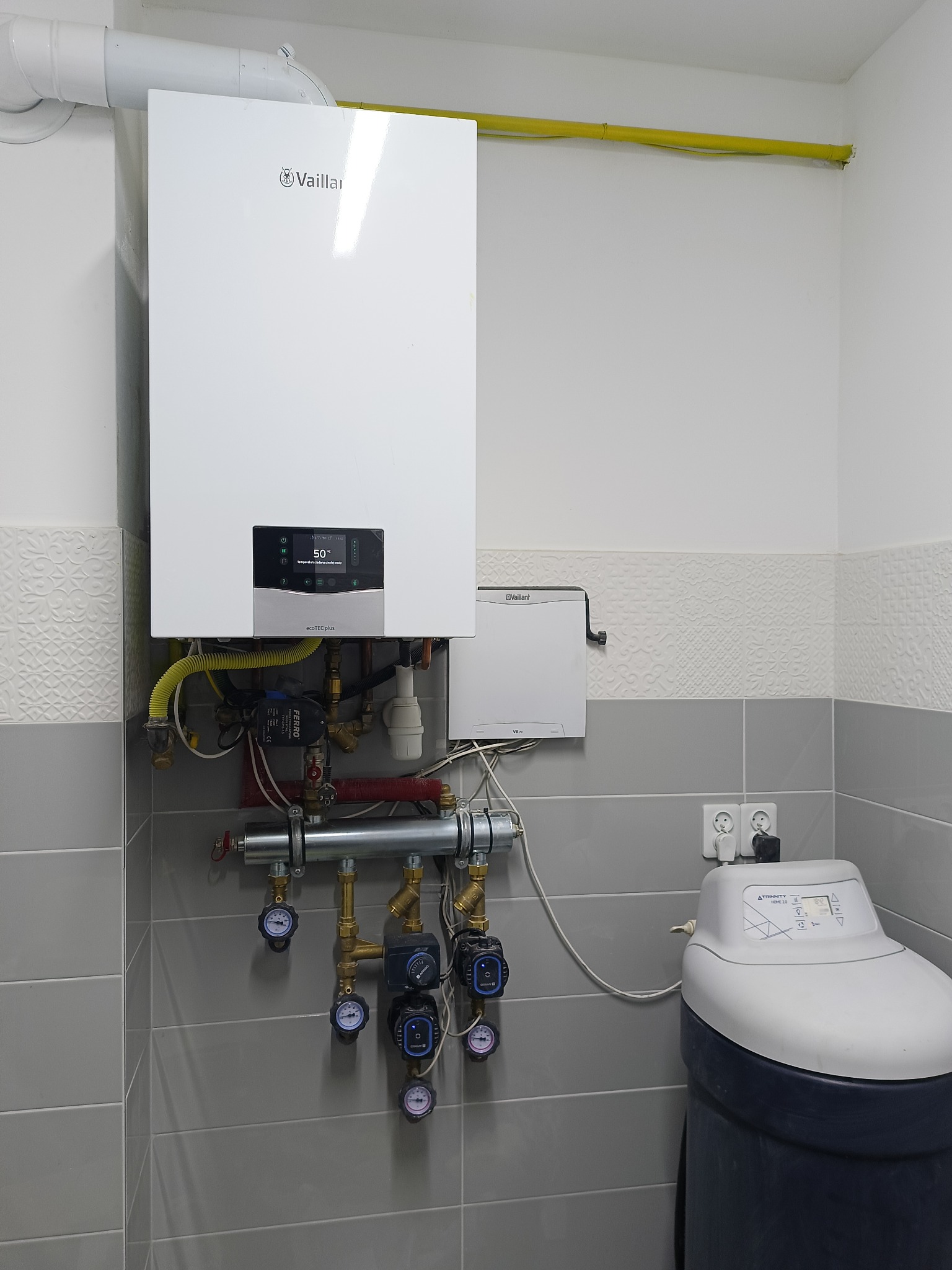 Biały piec gazowy Vaillant z żółtą rurą gazową, widoczne przyłącza hydrauliczne, manometry oraz zmiękczacz wody Trinity Home 2.0 na tle szarych płytek.