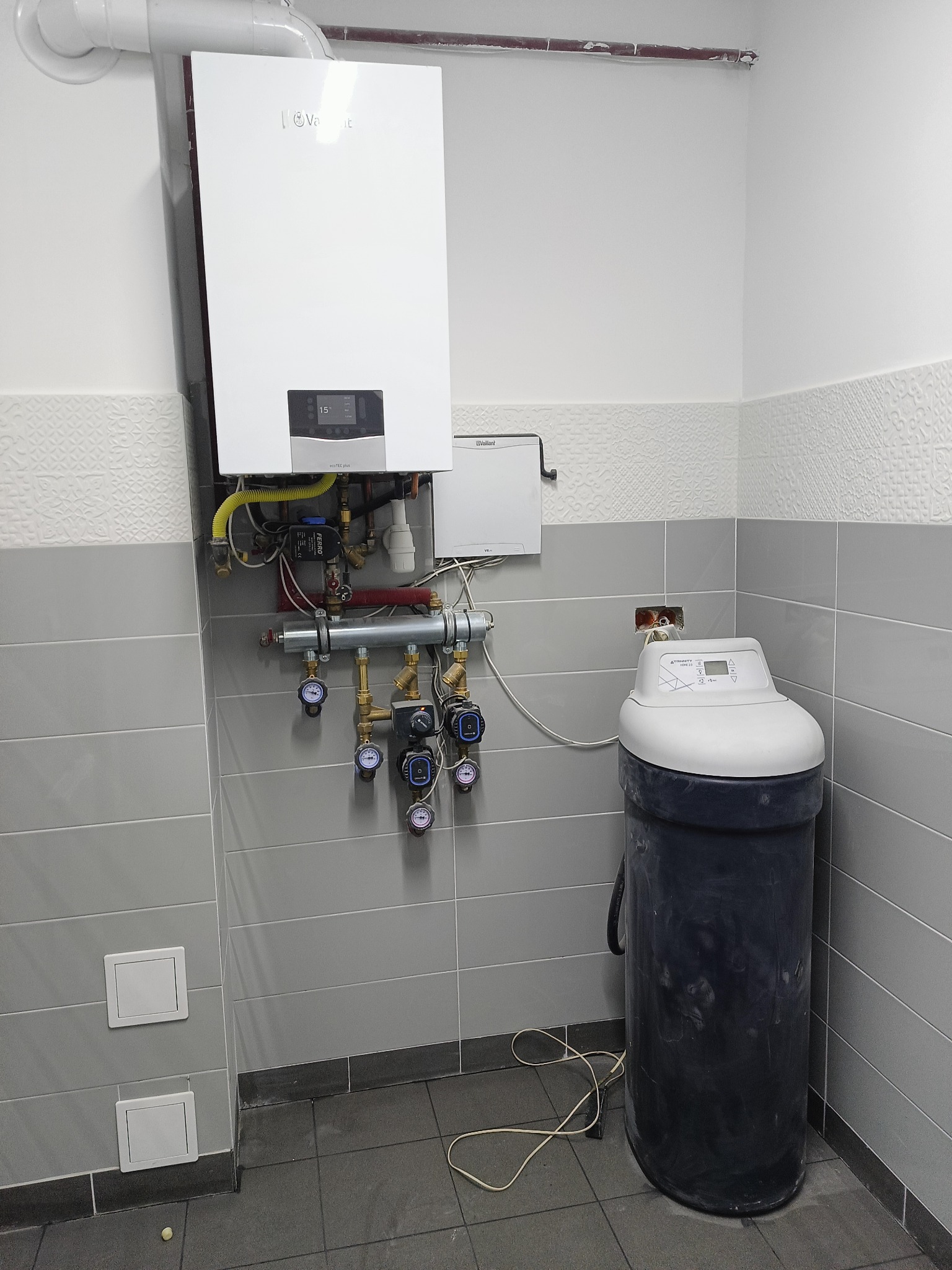 Biały piec gazowy Vaillant zamontowany na szarej, kafelkowej ścianie w Prusach, widoczne rury, manometry i zmiękczacz wody, całość w małej kotłowni.