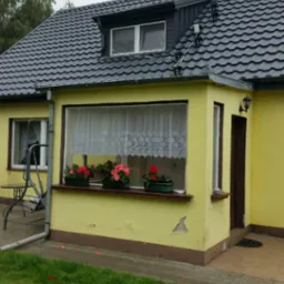 Żółty dom jednorodzinny z nowym, ciemnoszarym dachem z dachówki ceramicznej, widoczne zabezpieczenia przeciwśniegowe na połaci dachu, kwiaty w donicach na parapecie okna.